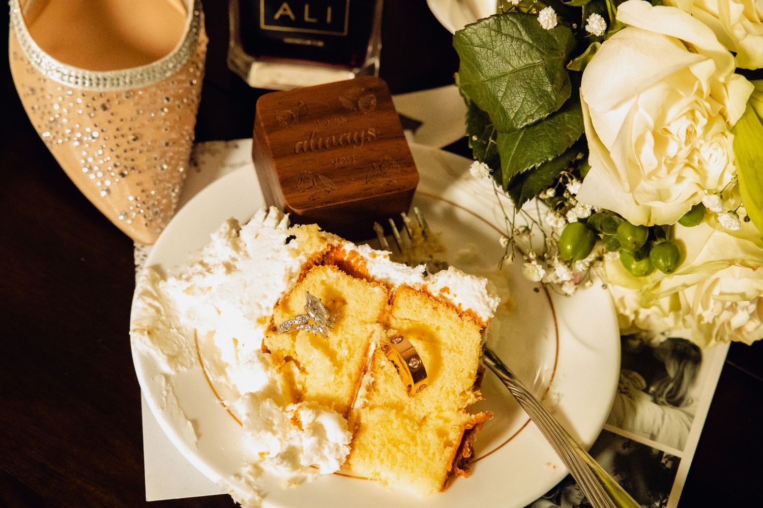 Two wedding rings resting on a slice of cake on a white plate beside white roses, a sparkly bridal shoe, and a wooden ring box.