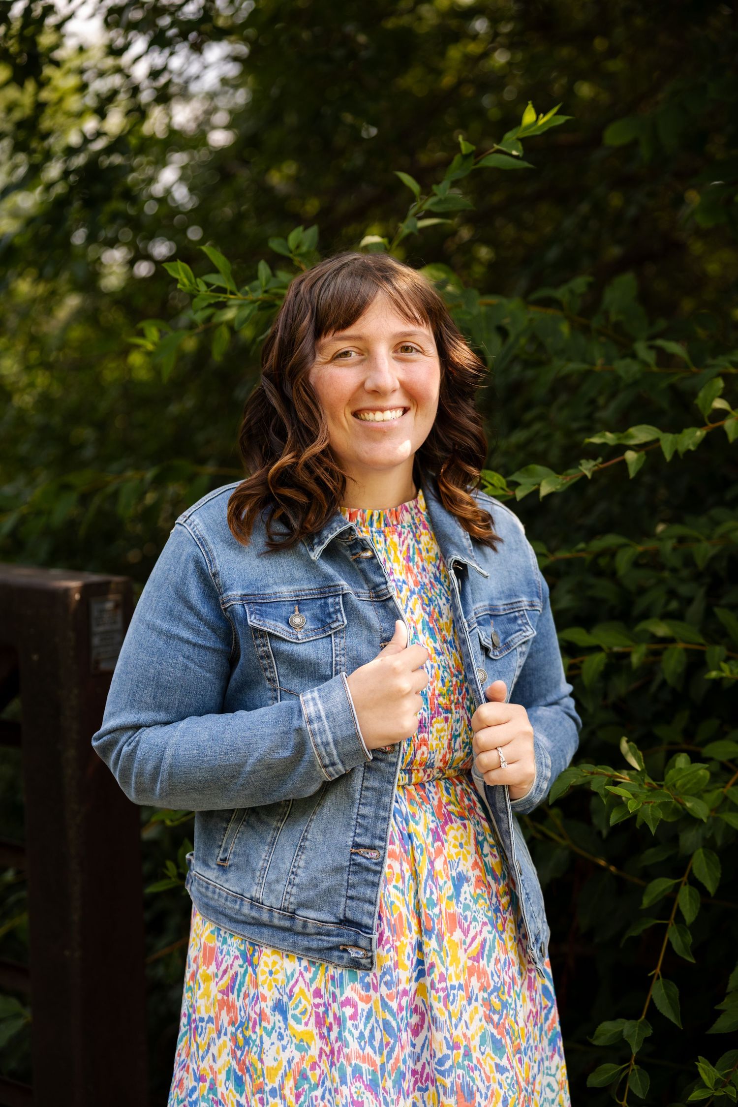 Smiling woman in denim jacket and floral dress standing outdoors among green foliage.