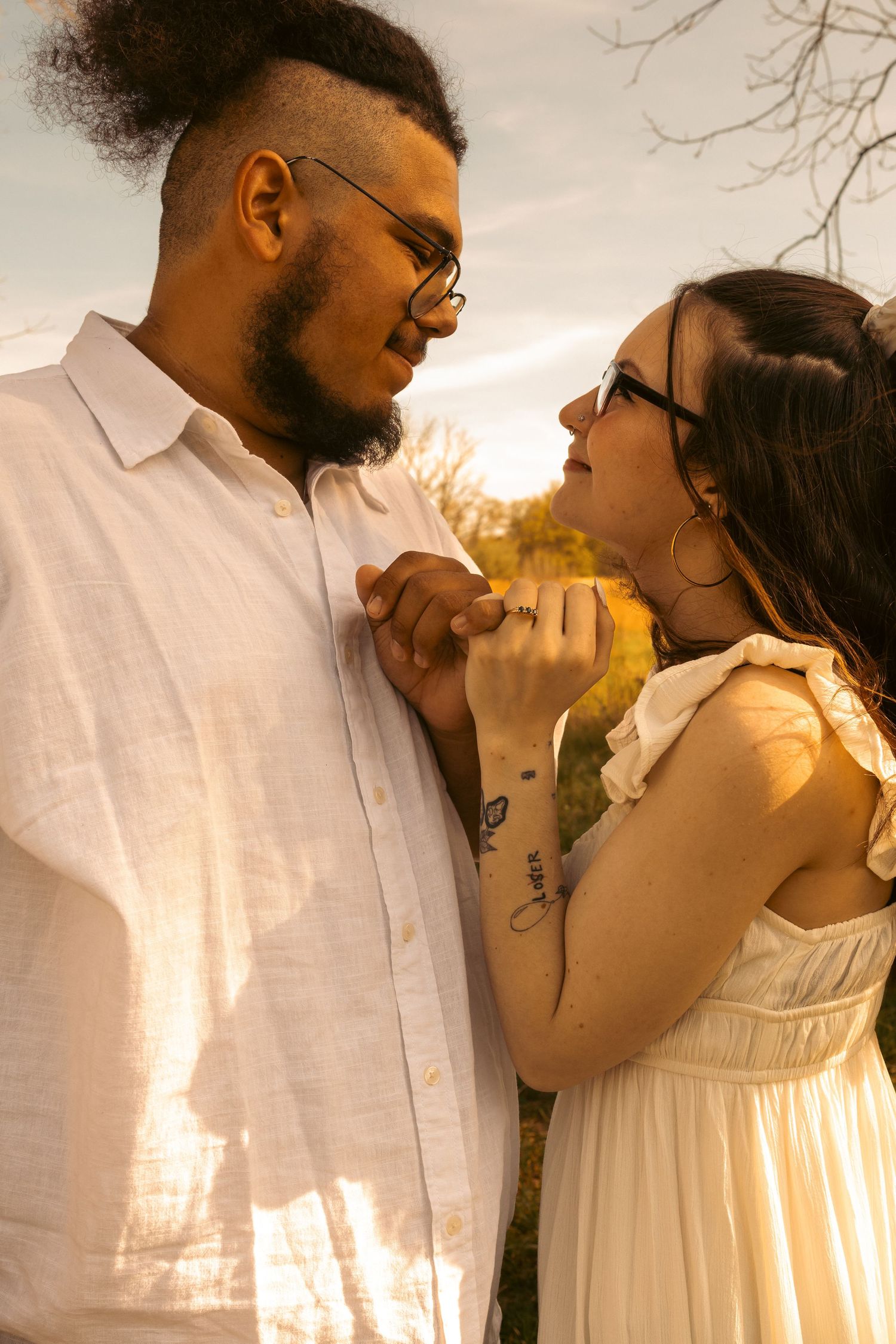 Interracial couple holding hands at golden sunset, man in white shirt and woman in white dress smiling.