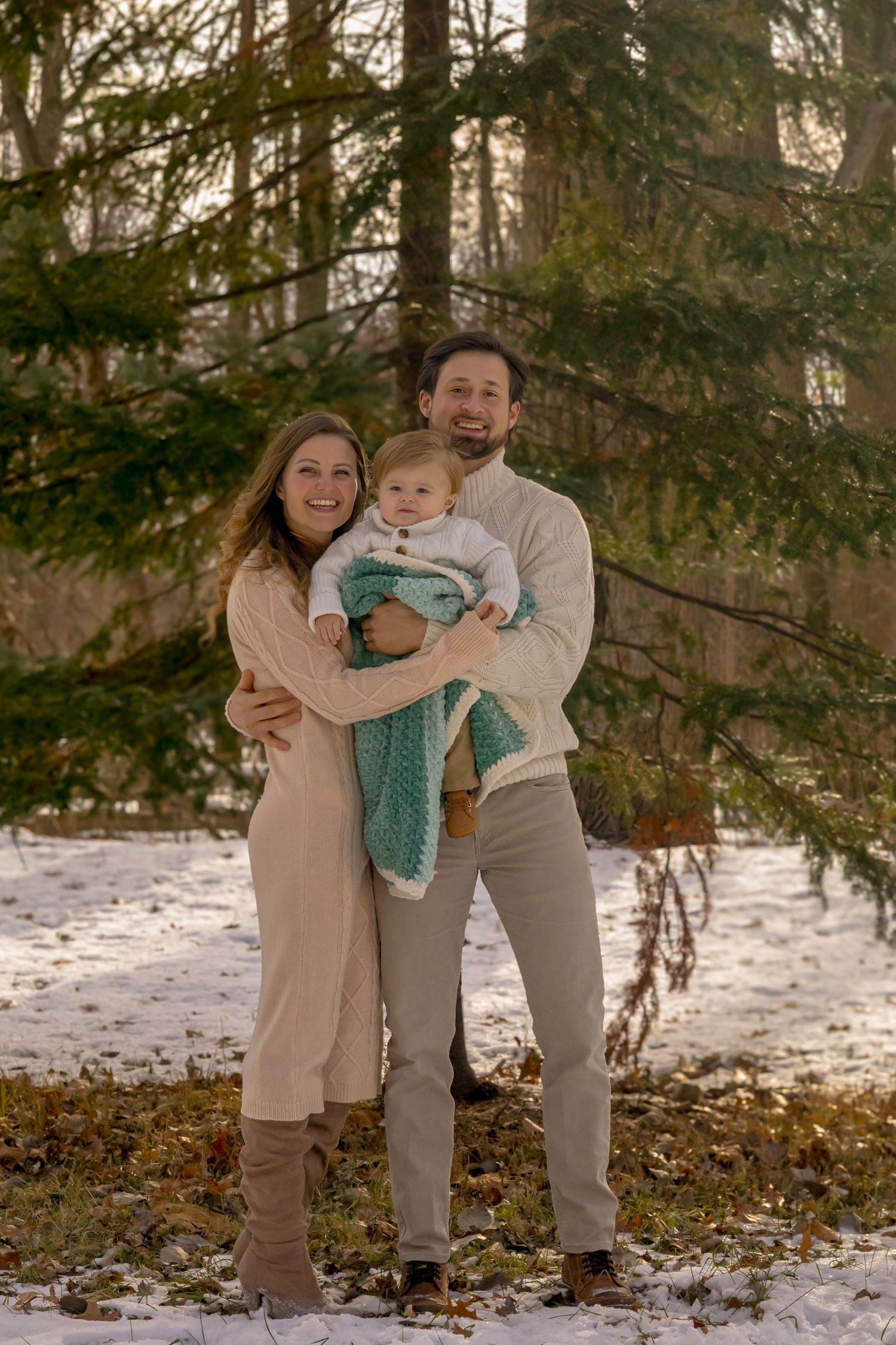 Happy family of three posing outdoors in winter, parents holding baby wrapped in teal blanket with snowy forest backdrop.