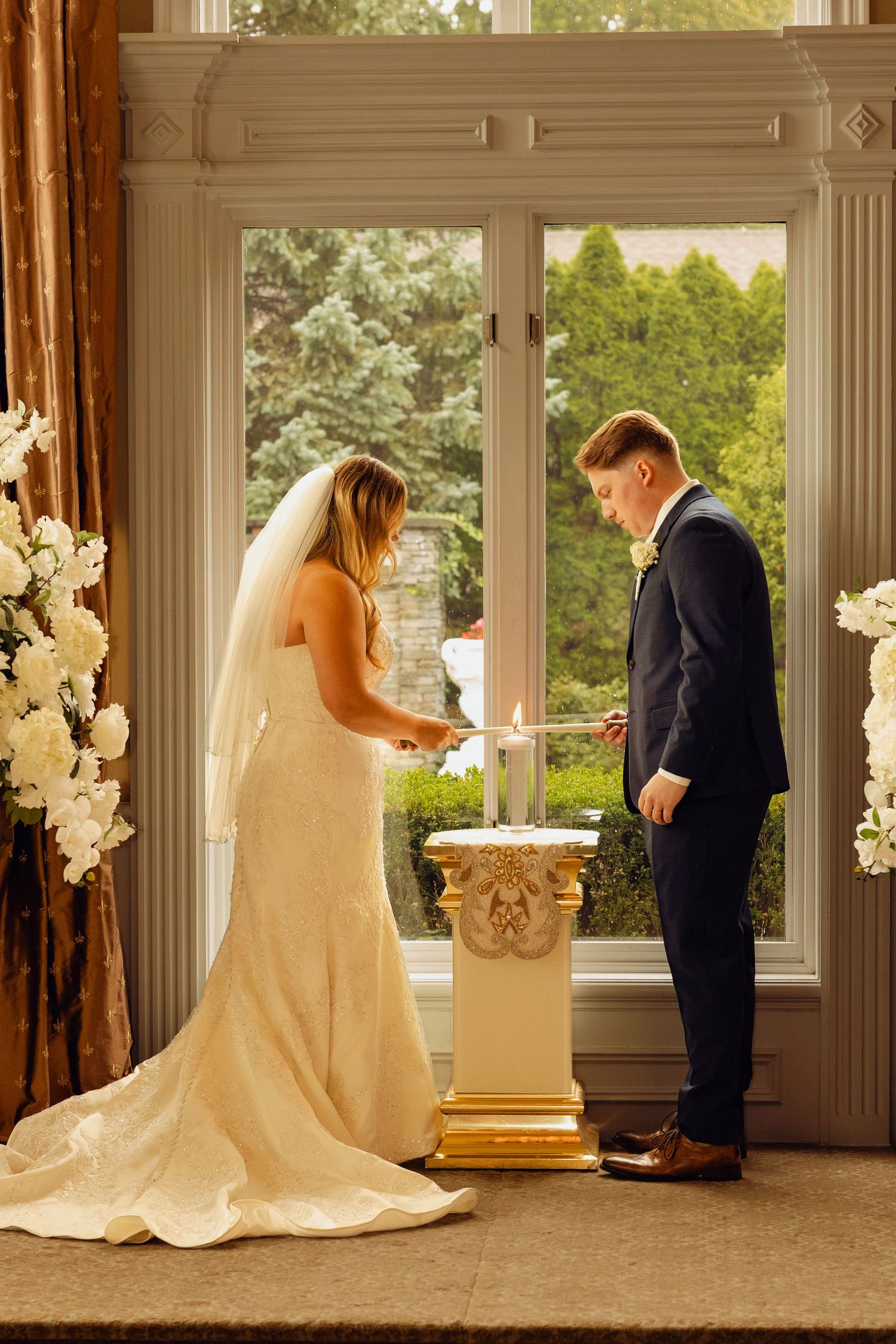 Bride in a white lace gown and groom in a navy suit light a unity candle on a gold pedestal beside a window during their wedding ceremony.