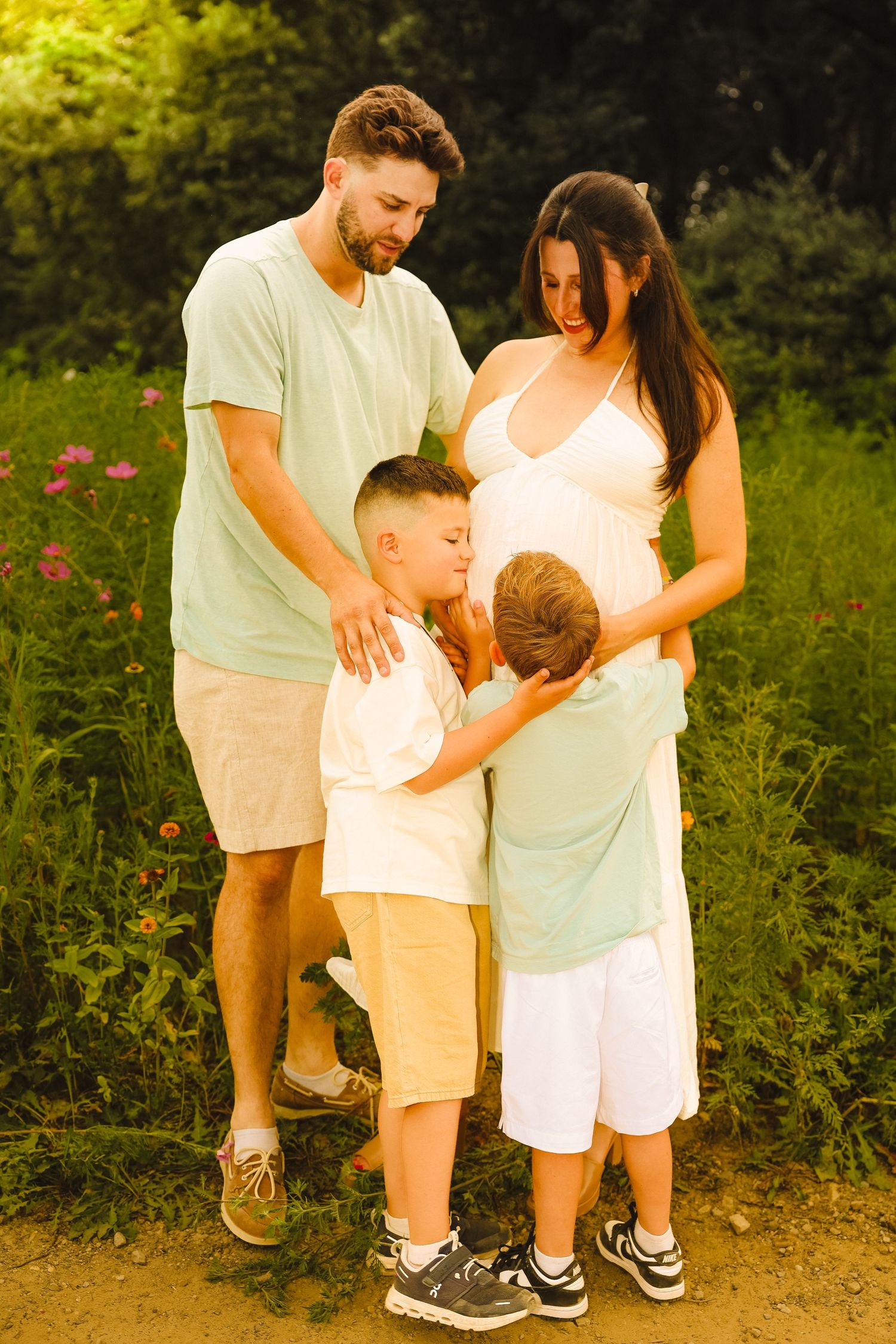 A pregnant woman and man stand outdoors with two young boys hugging her belly in a sunny garden setting.