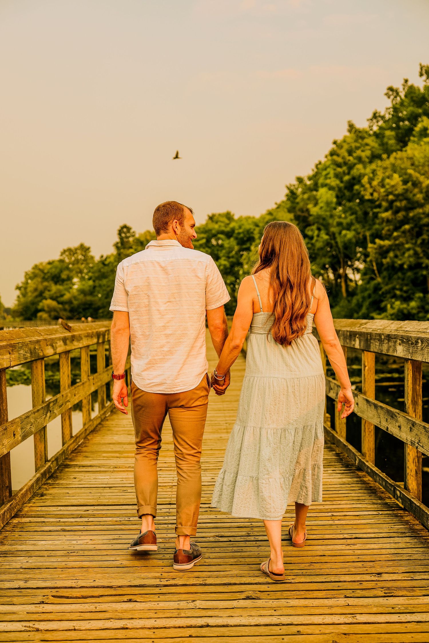 Couple holding hands walking on a wooden boardwalk surrounded by trees during golden hour sunset.