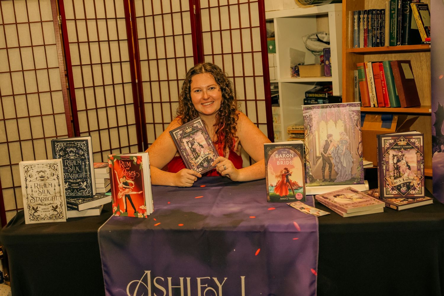 Author Ashley J smiles at a book signing table displaying fantasy novels in a cozy bookstore setting.