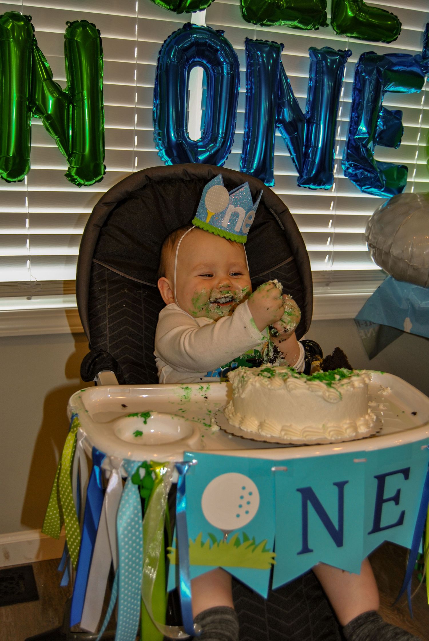A birthday cake with white frosting and blue decorations sits on a table beneath balloon letters and a festive banner.