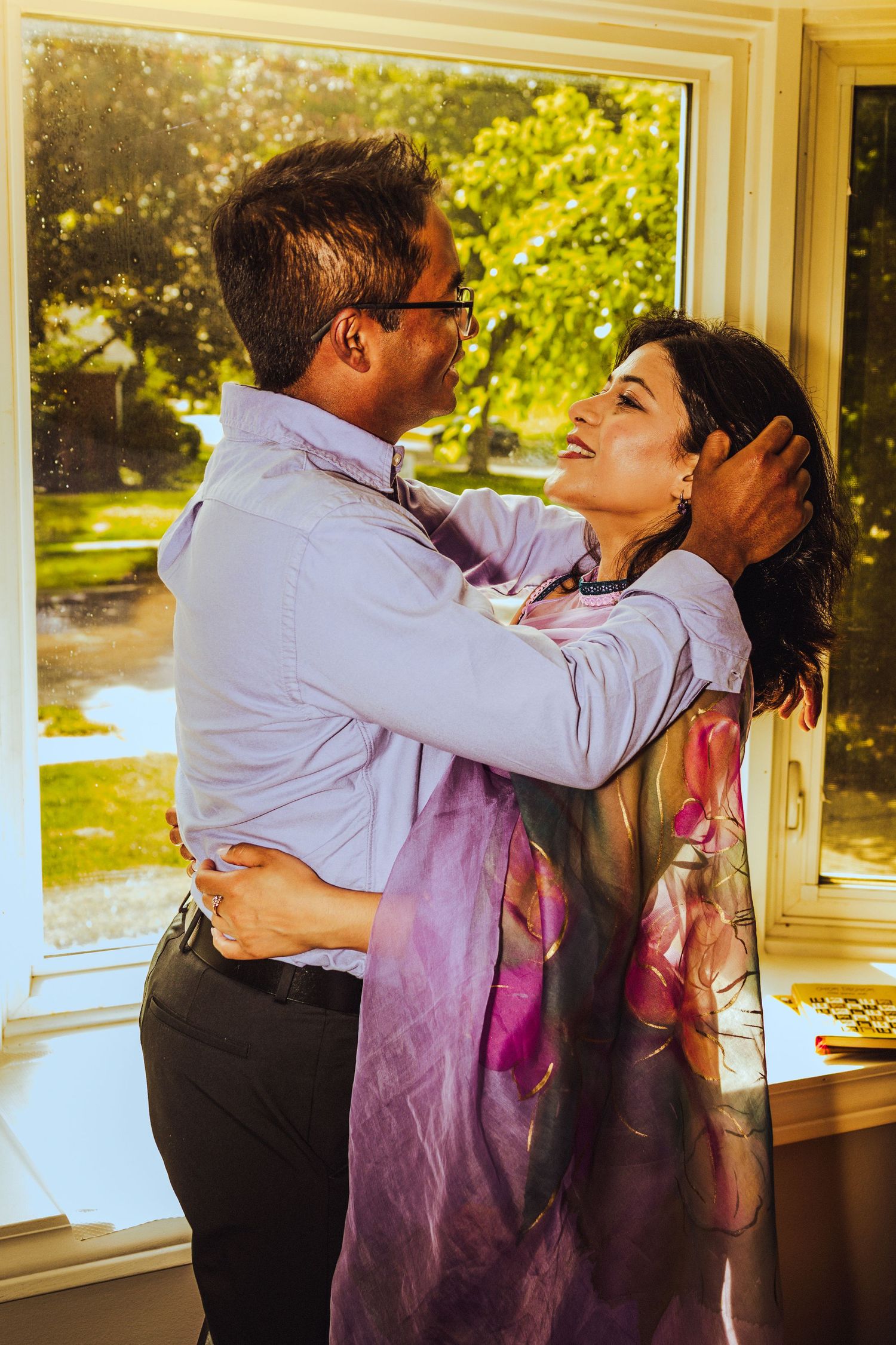 A couple embraces warmly near a sunlit window, the woman in a floral scarf smiling up at the man in glasses.