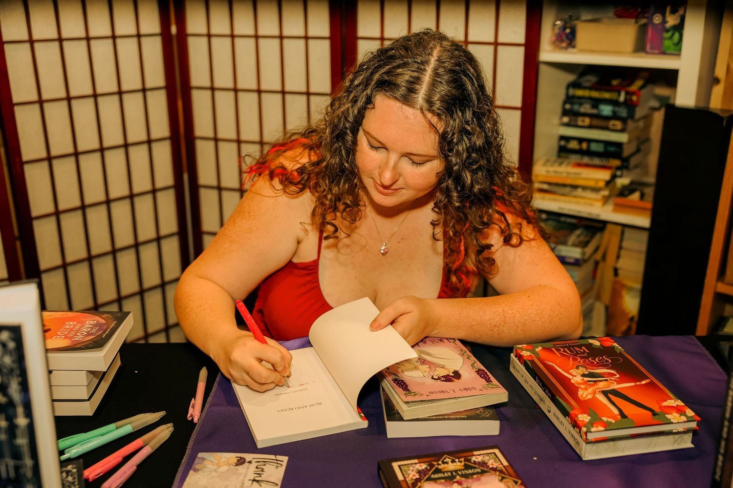 Author signing books at a bookstore table surrounded by stacked novels during a book signing event.