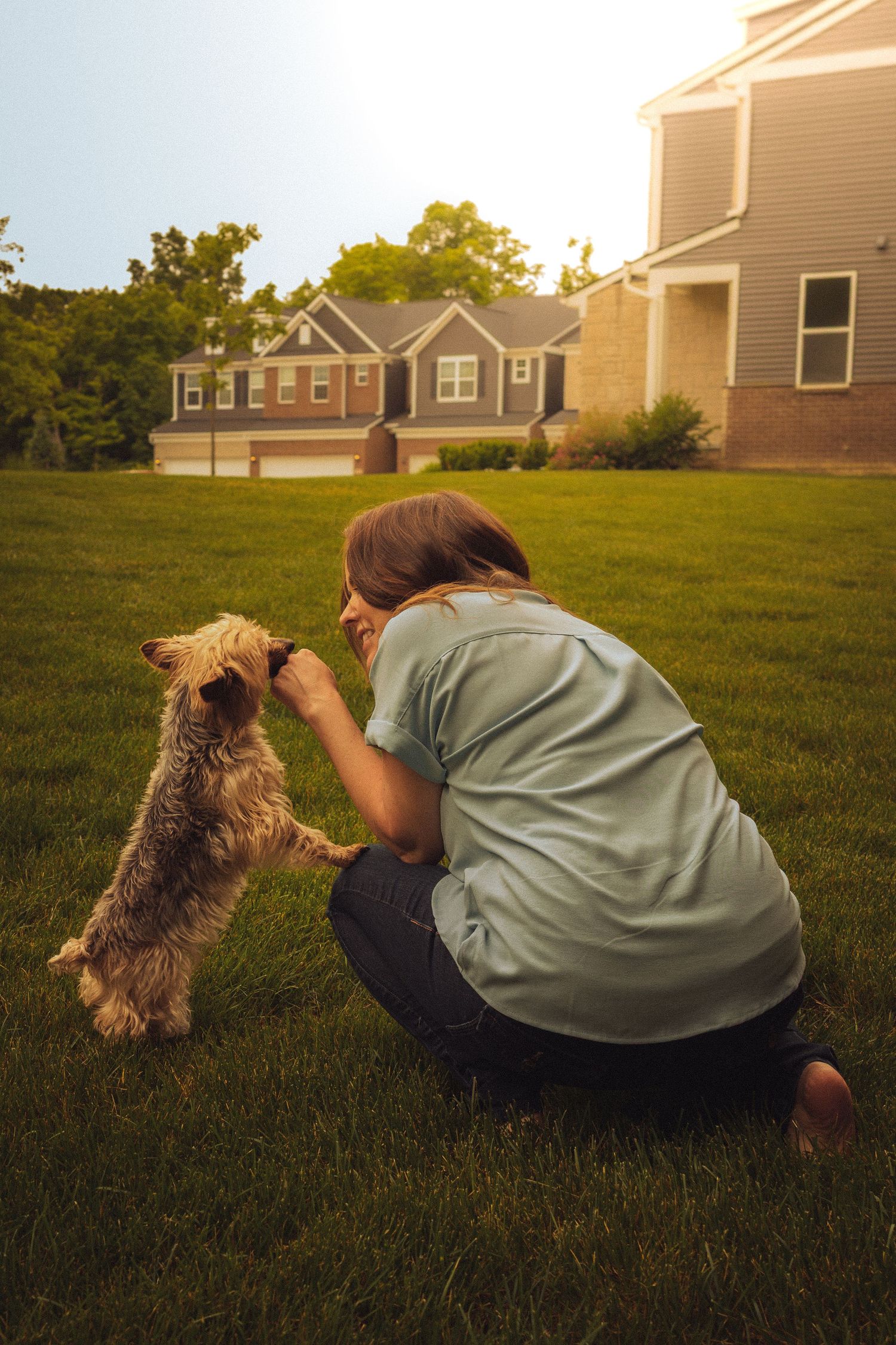 Woman in teal shirt crouching on green lawn, petting small terrier dog near suburban homes at golden hour.