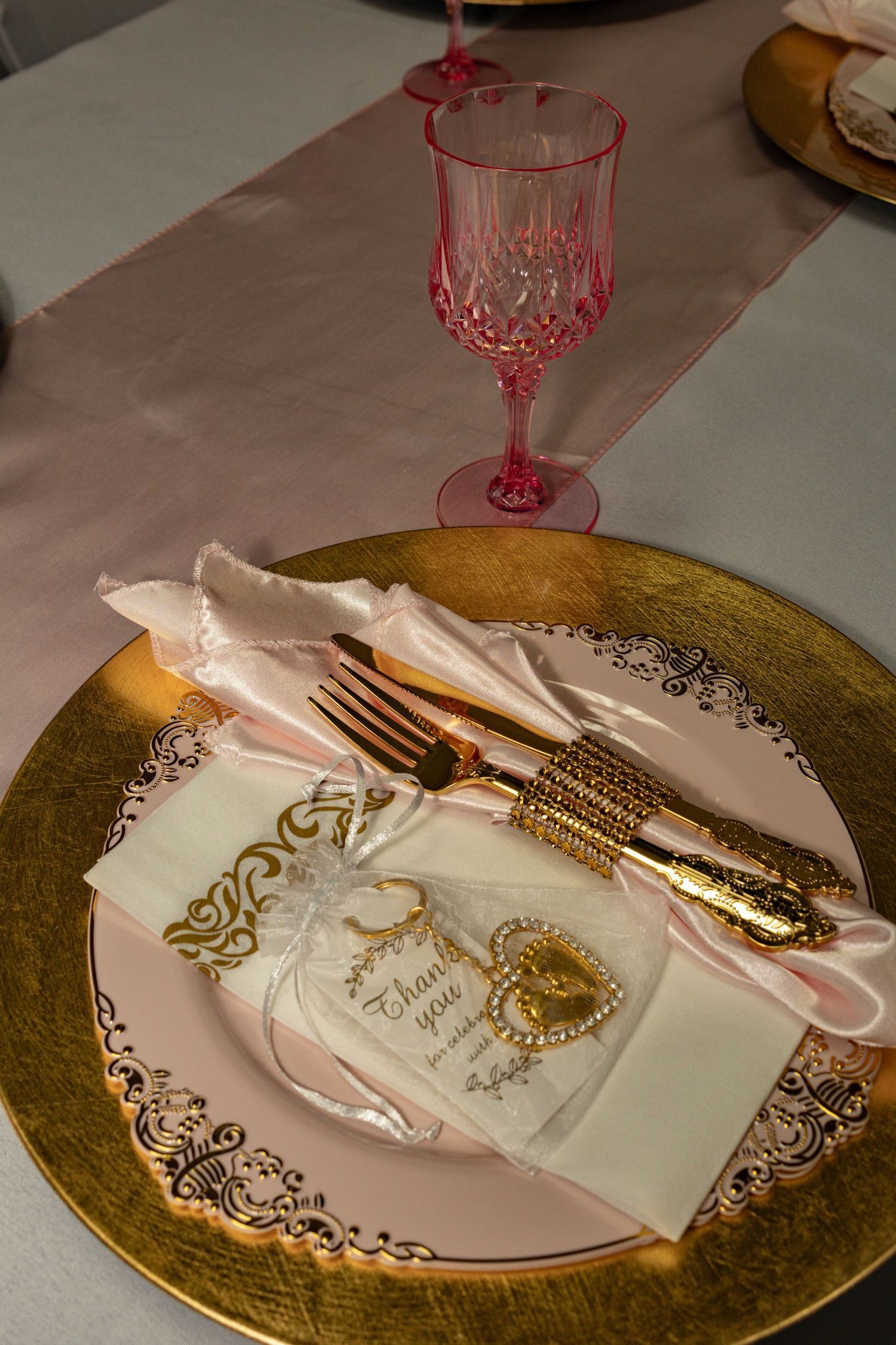Elegant gold charger plate with pink napkin, gold cutlery, thank you favor, and pink crystal wine glass on a formal table.