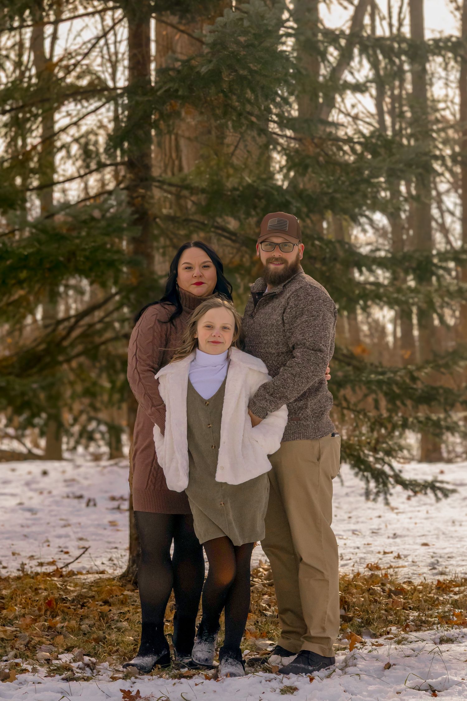 Family of three posing outdoors in snowy winter forest, wearing cozy fall outfits among tall evergreen trees.