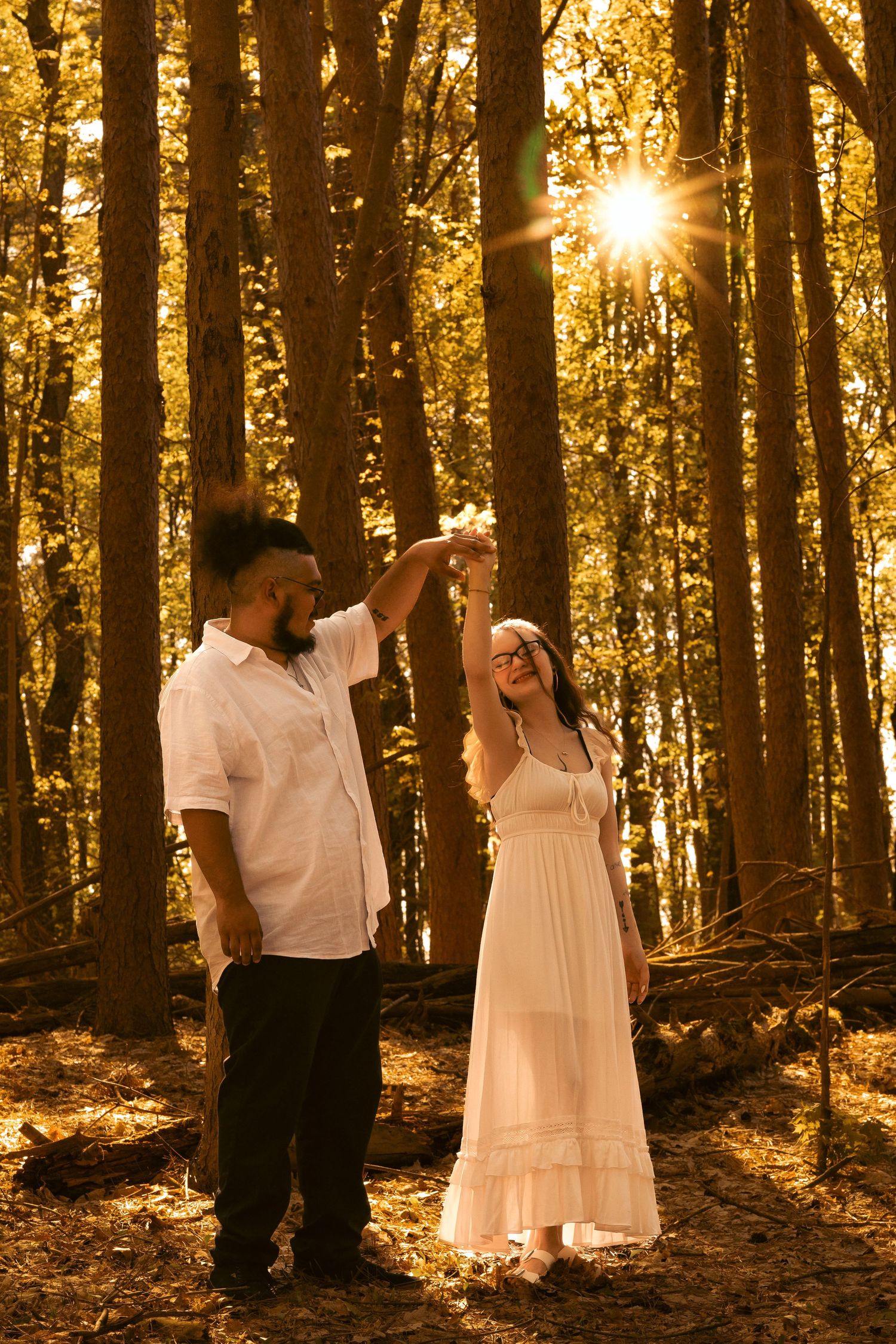 Couple dancing in golden sunlit forest, woman in white dress, man in white shirt, surrounded by tall trees.