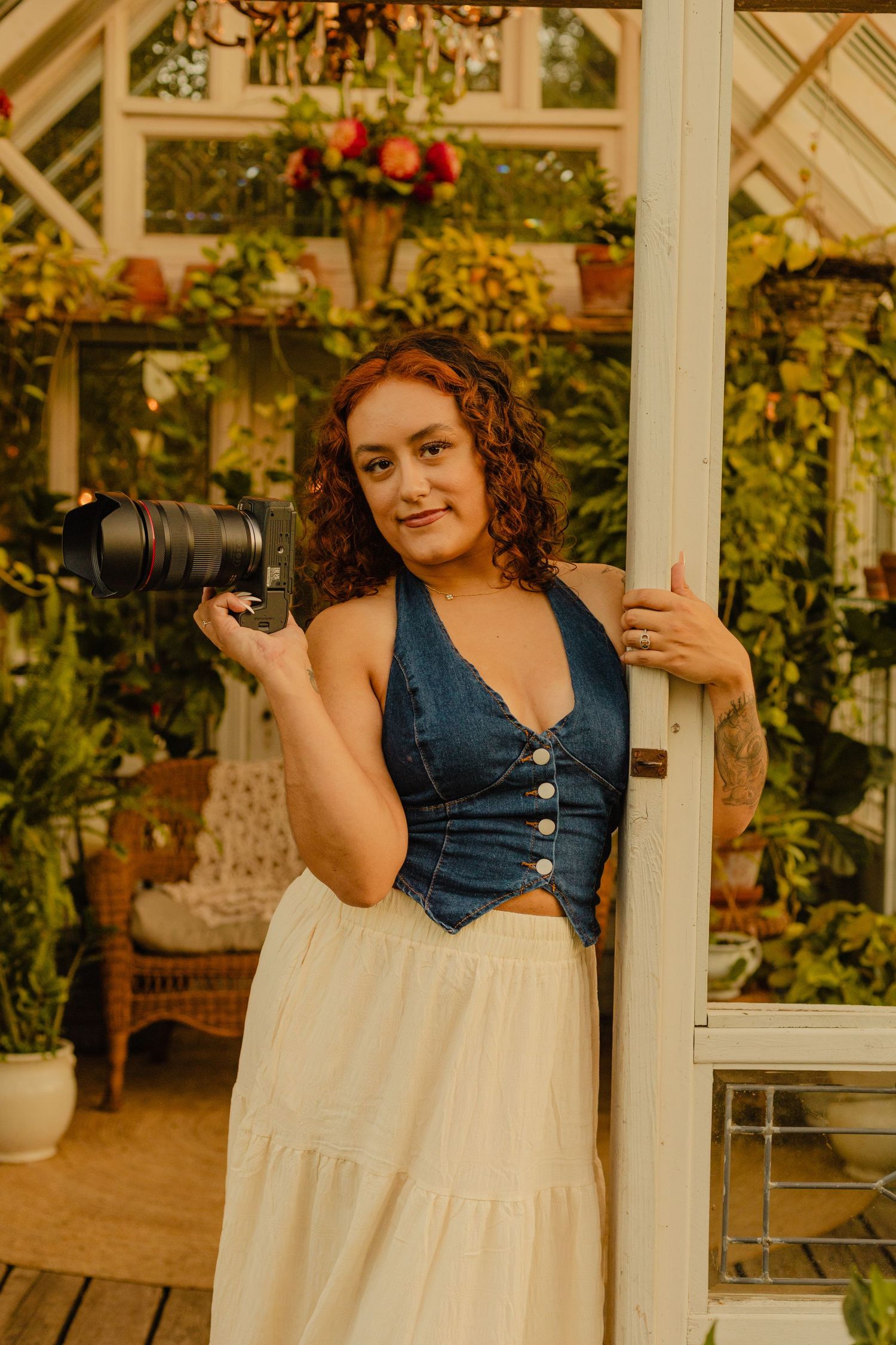 A young woman with curly red hair holds a camera in a lush greenhouse, wearing a denim top and white skirt.