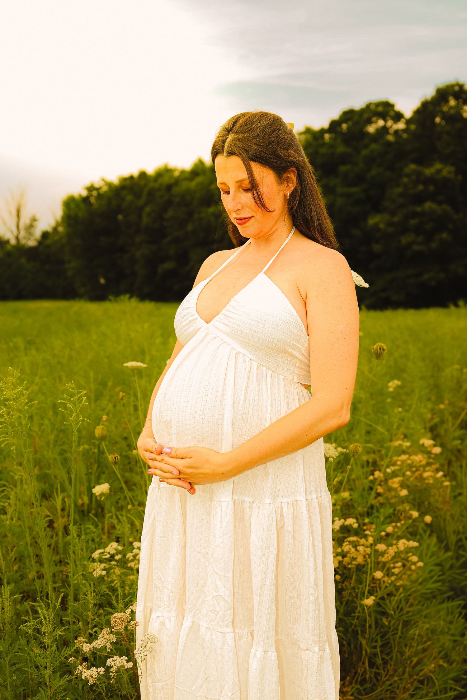 Pregnant woman in white halter dress cradling her baby bump in a sunny green meadow with wildflowers.