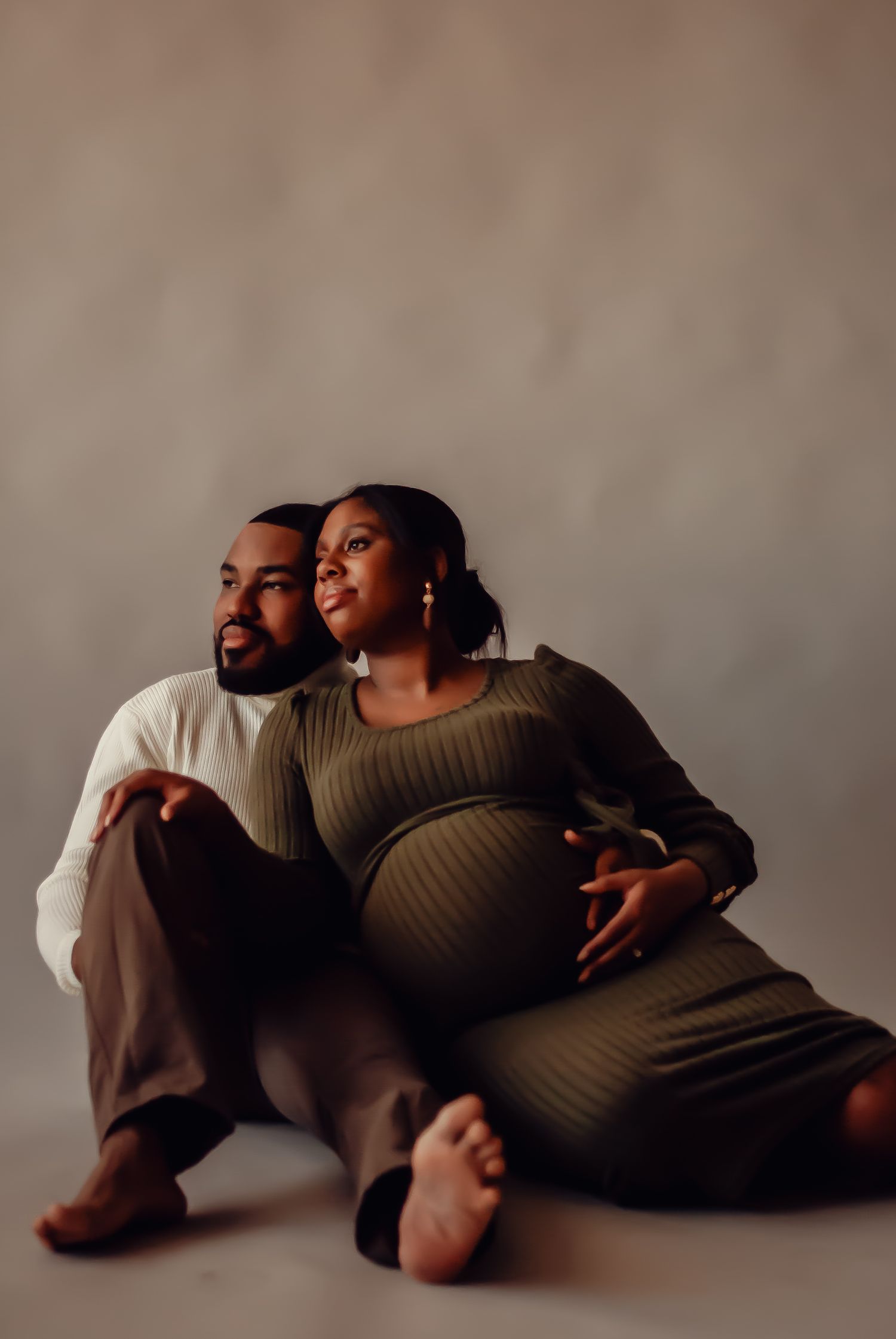 Couple sits together on floor during intimate maternity photo session in olive green dress and brown pants.