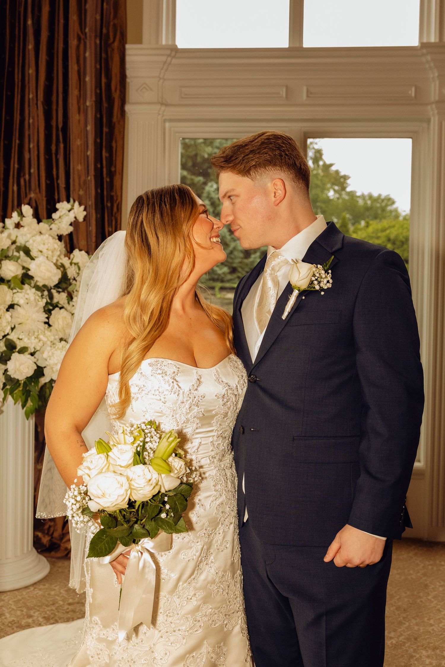 Bride in a lace strapless gown holding a white rose bouquet shares a close, tender moment with the groom in a navy suit at a wedding.