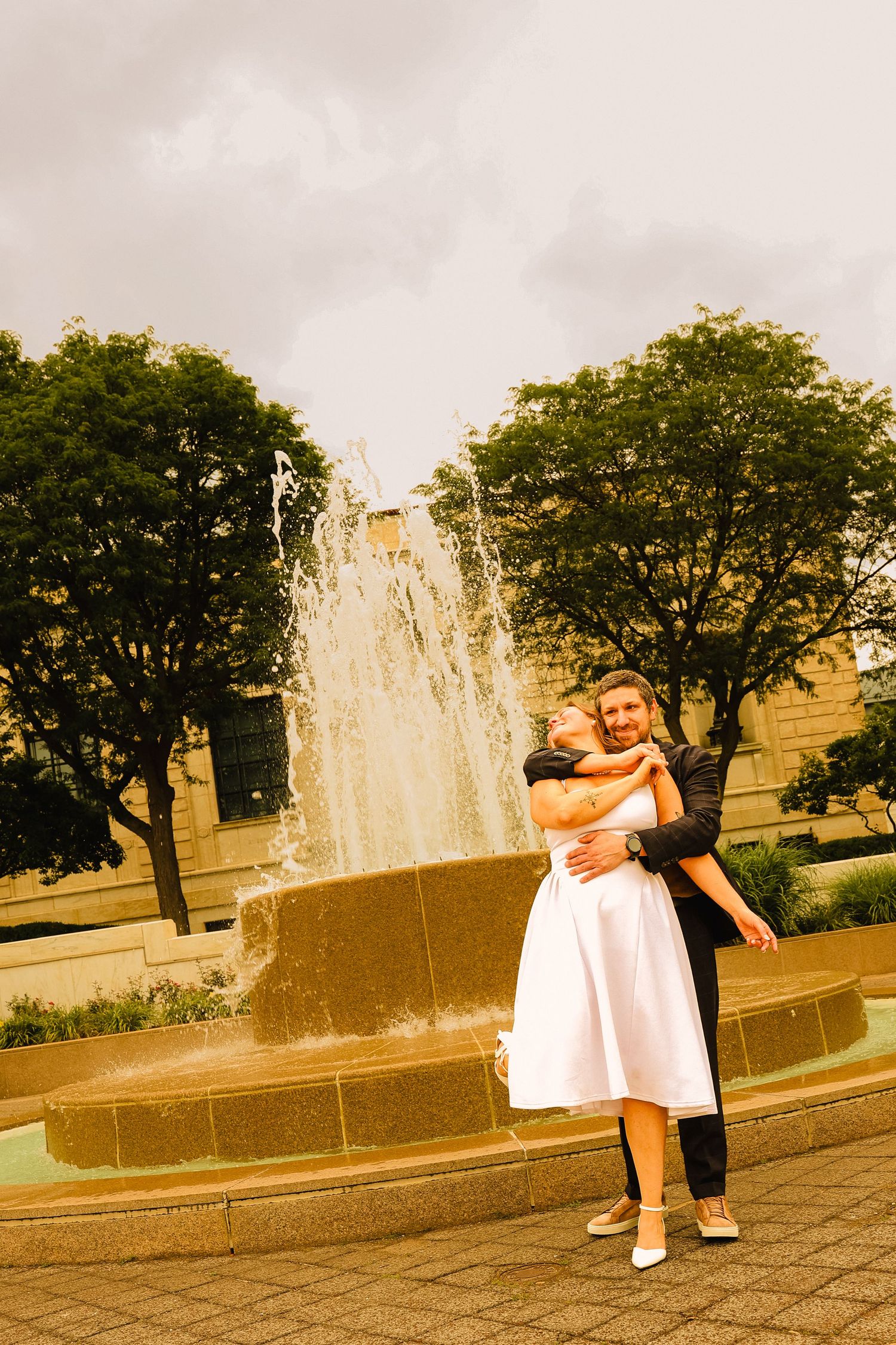 Couple embracing and smiling in front of a fountain outdoors, with trees and cloudy sky in the background.