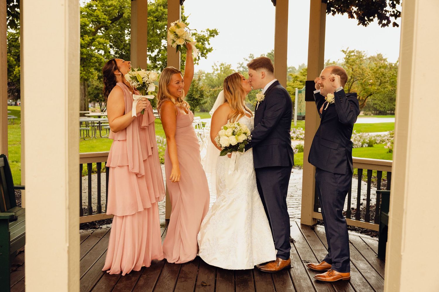 Bride in white gown kisses groom on a wooden gazebo while bridesmaids in blush dresses and a groomsman cheer with bouquets.