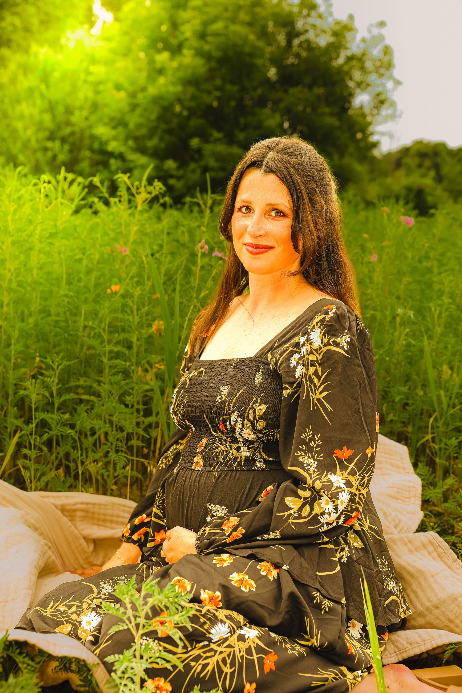 A woman in a floral black dress sits gracefully in a lush green field, bathed in warm golden sunlight.