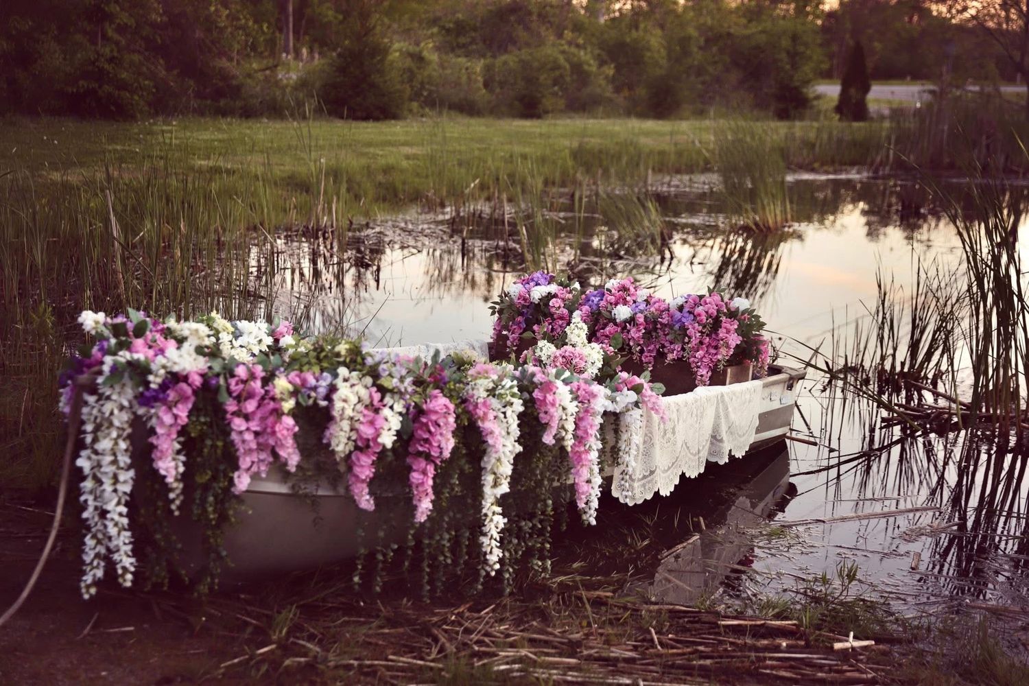 A flower-adorned rowboat floating on a calm lake surrounded by lush green marsh grasses at dusk.