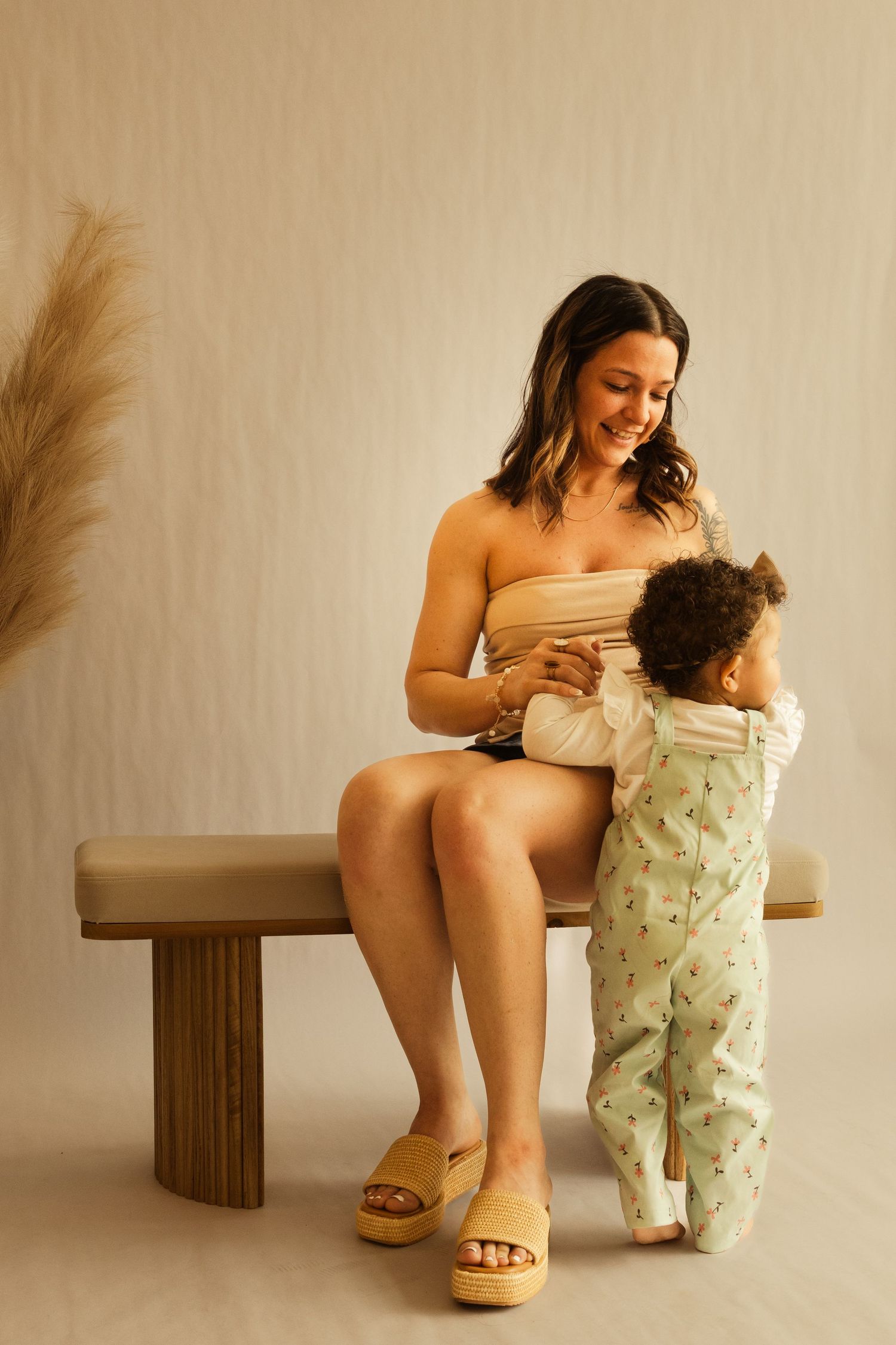 A smiling woman in a beige tube top sits on a bench while a toddler in green floral overalls stands beside her.