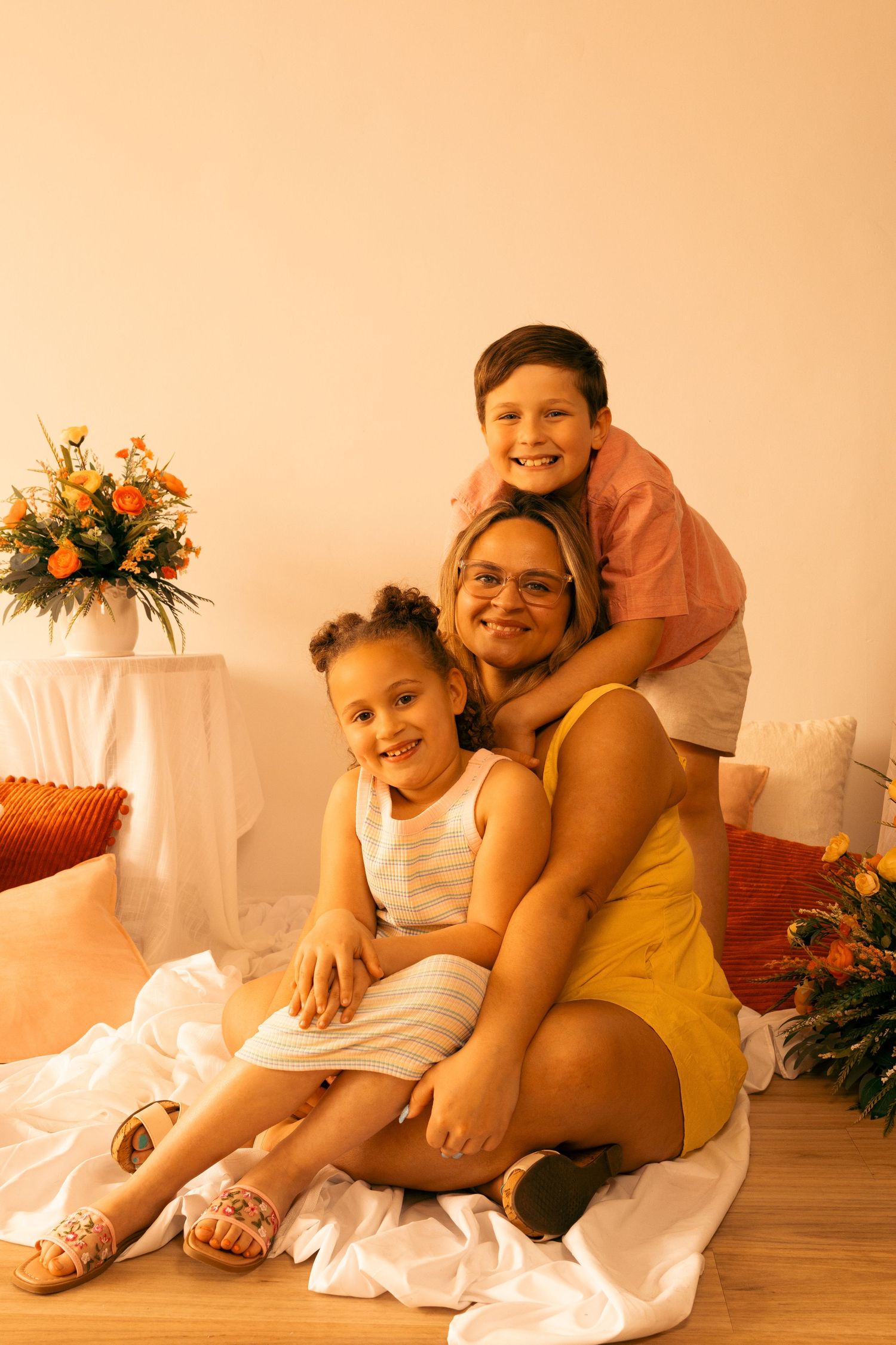 A woman in a yellow dress sits on the floor smiling with two young children in a warmly lit room with orange flowers.