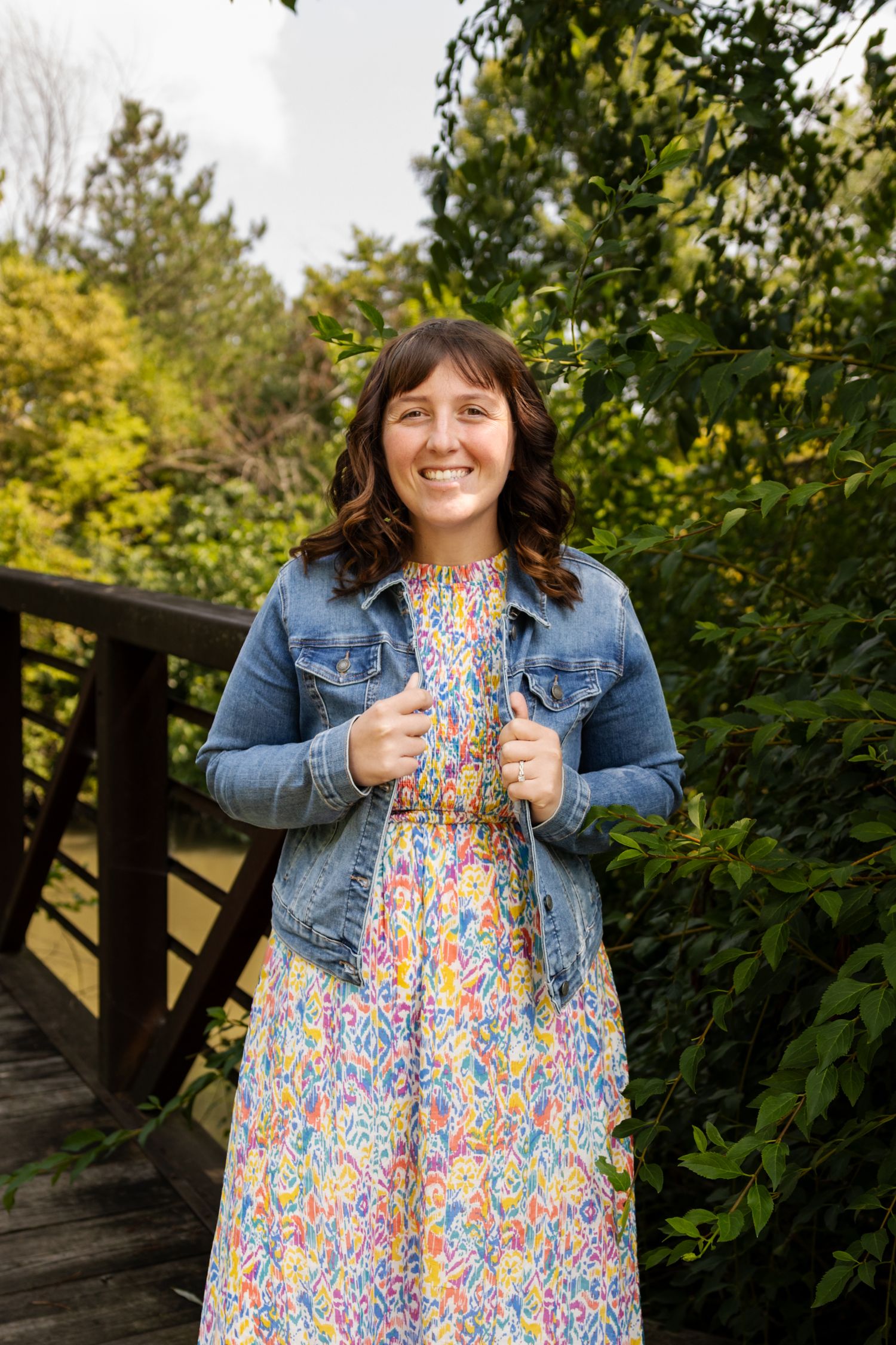 Smiling young woman in colorful floral dress and denim jacket standing on wooden bridge surrounded by green trees.