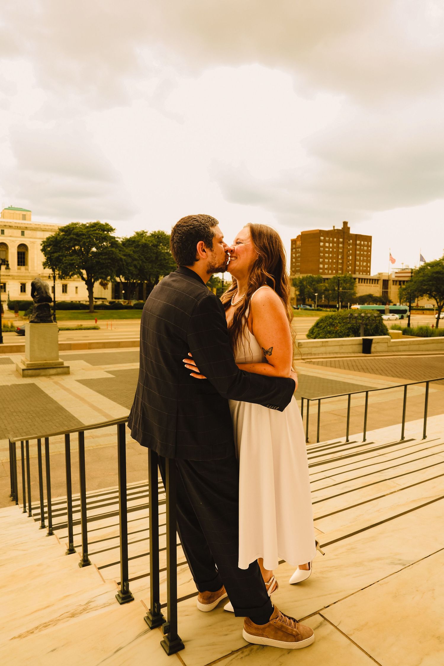 Couple sharing a romantic kiss on outdoor steps with a cloudy sky and city plaza in the background.