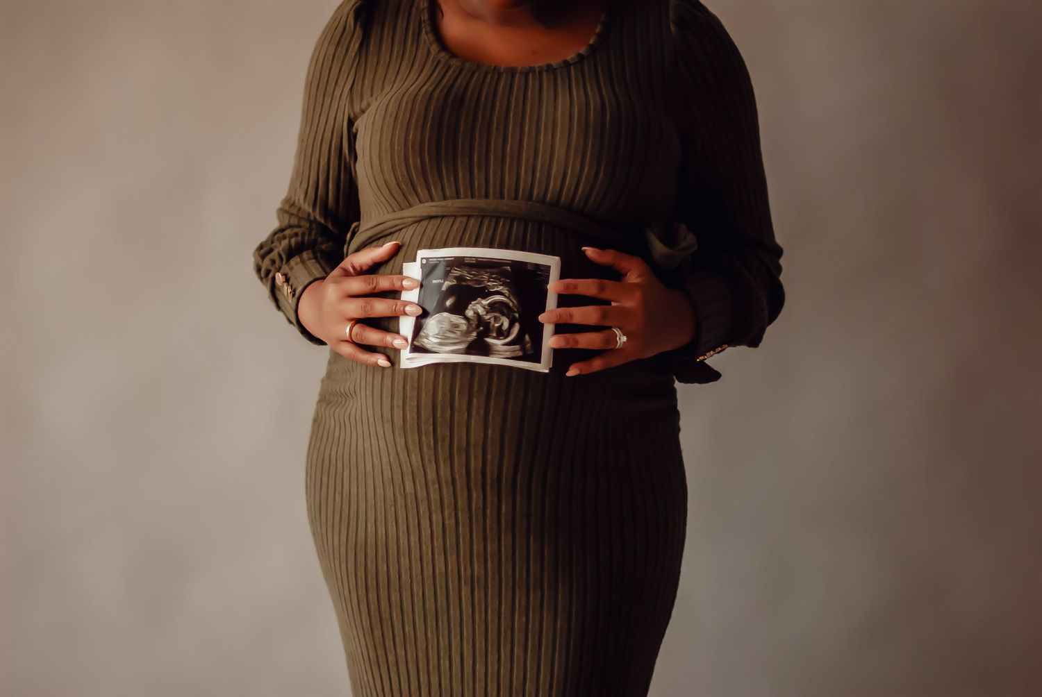 Someone in a brown ribbed dress holds an ultrasound photo against their pregnant belly in a minimalist maternity photo.