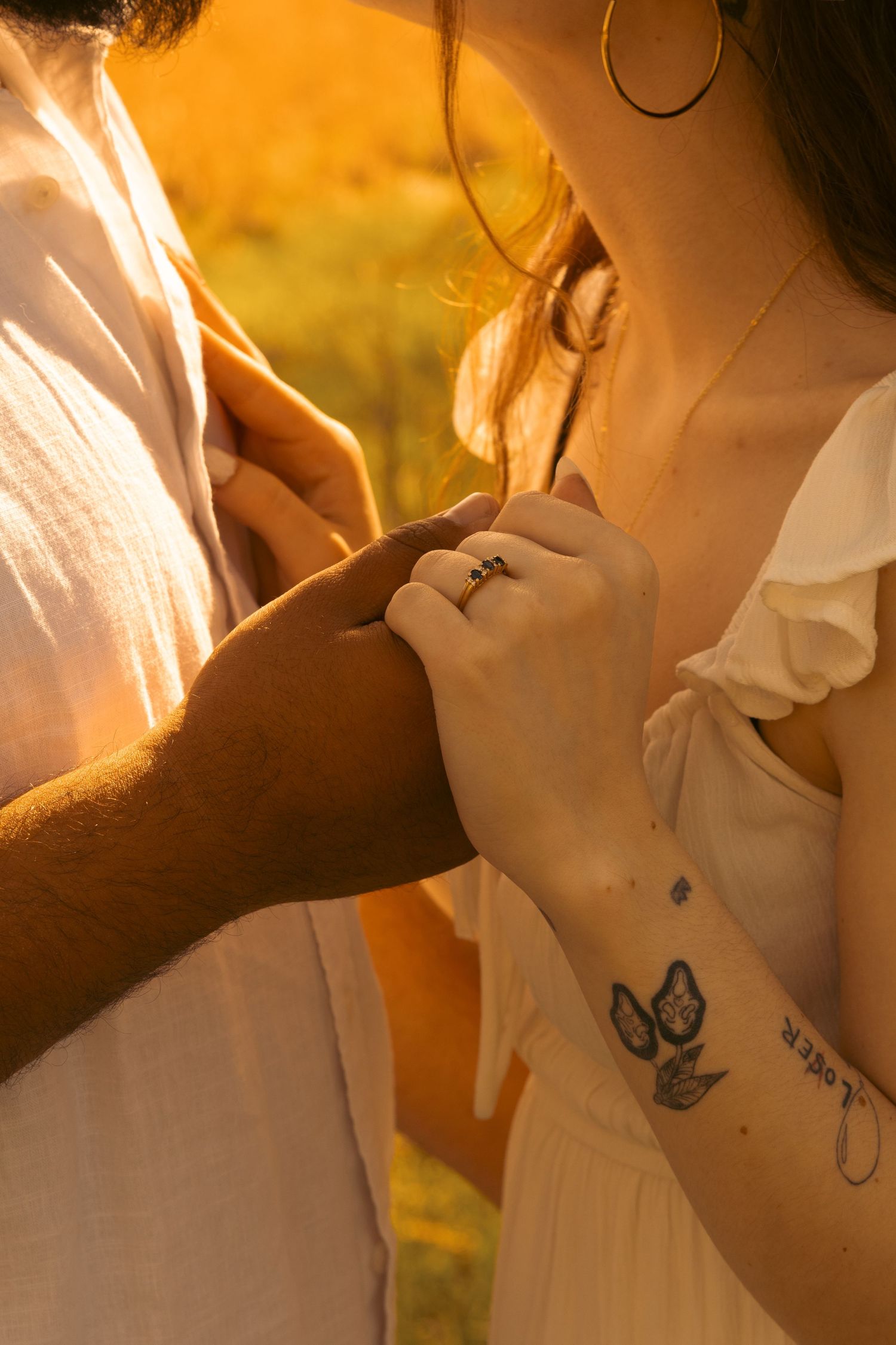 Couple holding hands at golden hour, woman wearing a ring and butterfly tattoo on her wrist, warm sunset light.