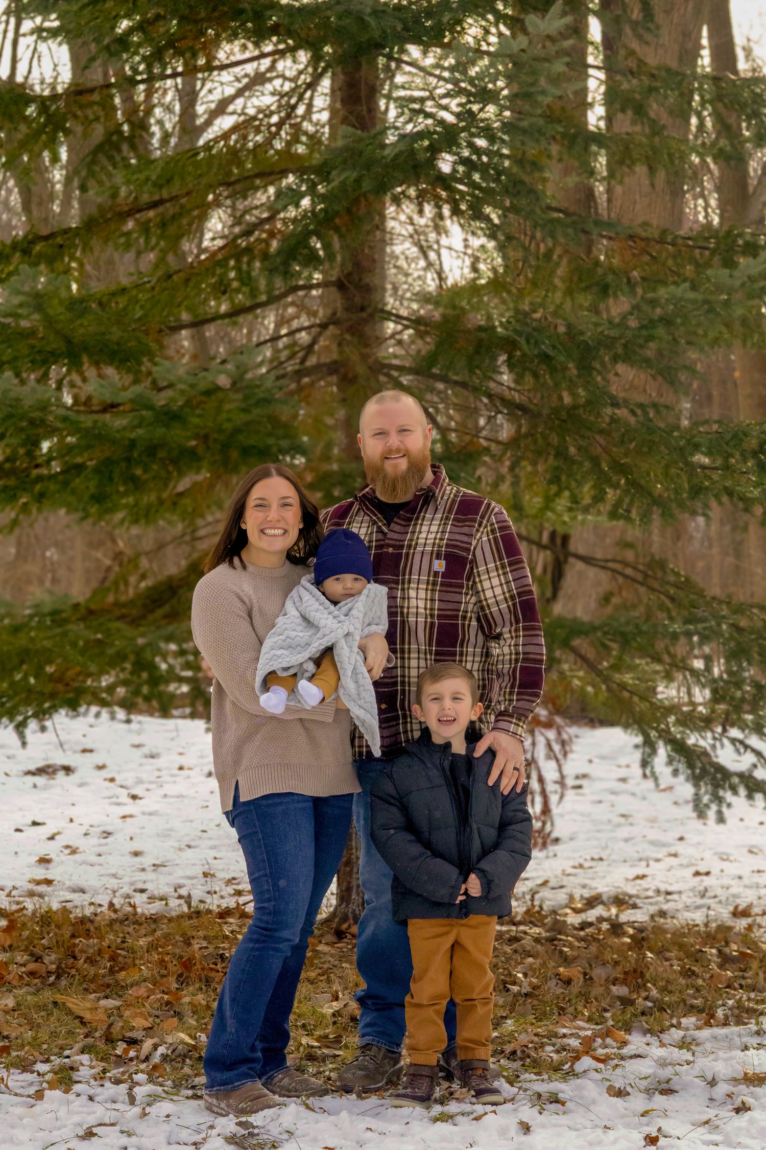 Family of four posing outdoors in winter, parents holding baby and young boy, evergreen trees and snow in background.