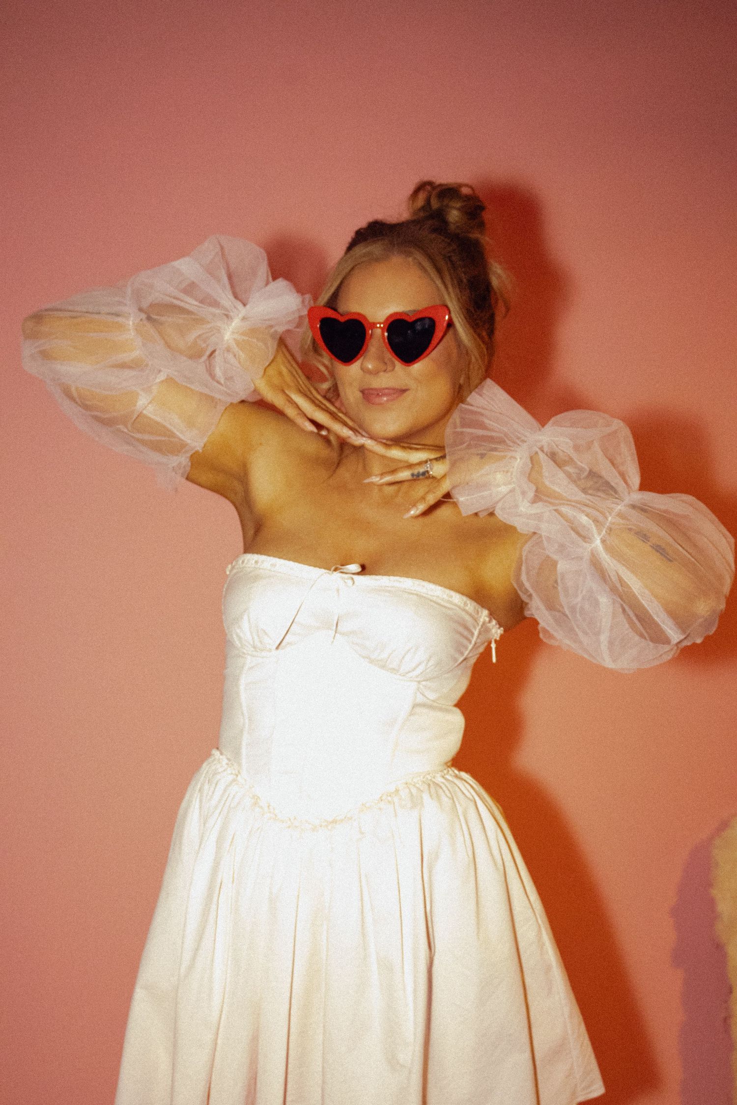 Woman in white strapless dress with tulle sleeves and red heart sunglasses posing against a pink background.