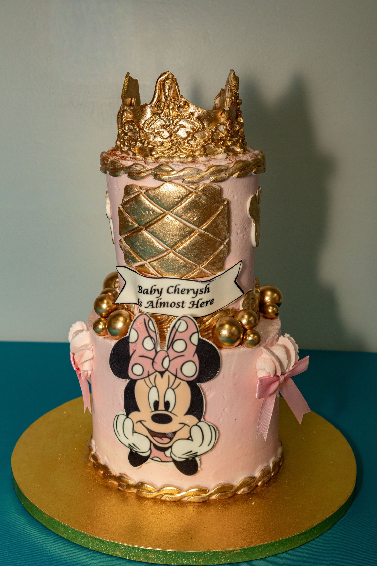 Two-tier pink and gold Minnie Mouse baby shower cake with crown topper reading Baby Cherysh Almost Here.
