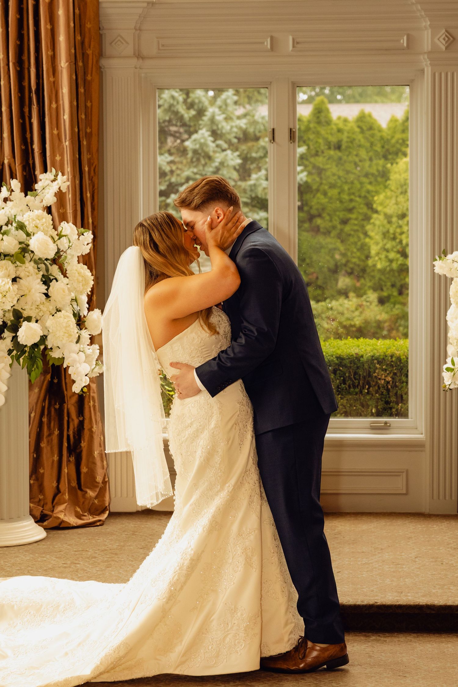 Bride in a white lace gown and veil kisses groom in a navy suit beside white floral arrangements during a wedding ceremony.