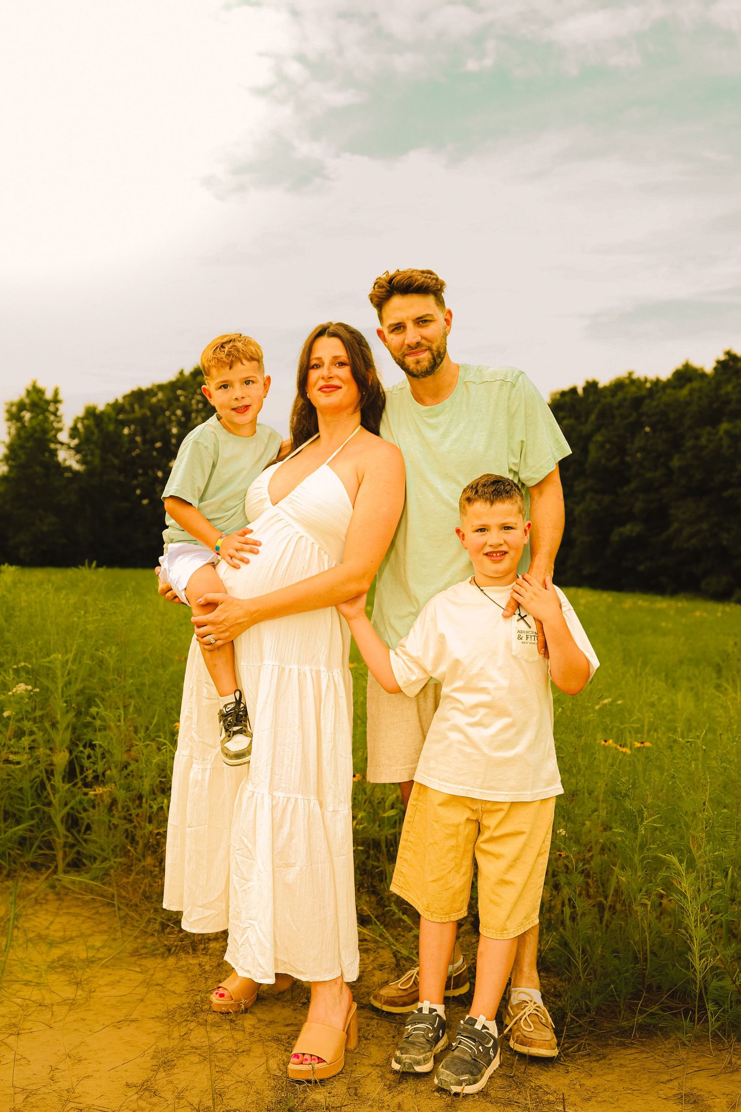 A happy pregnant family of four poses outdoors in a green field, with two young boys and their parents smiling warmly.