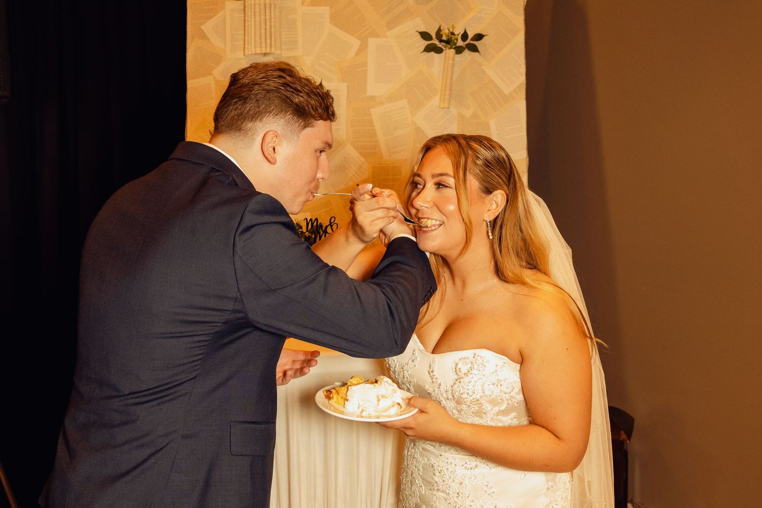 Groom in a dark suit feeds wedding cake to a smiling bride in a strapless white gown during their cake-cutting ceremony.