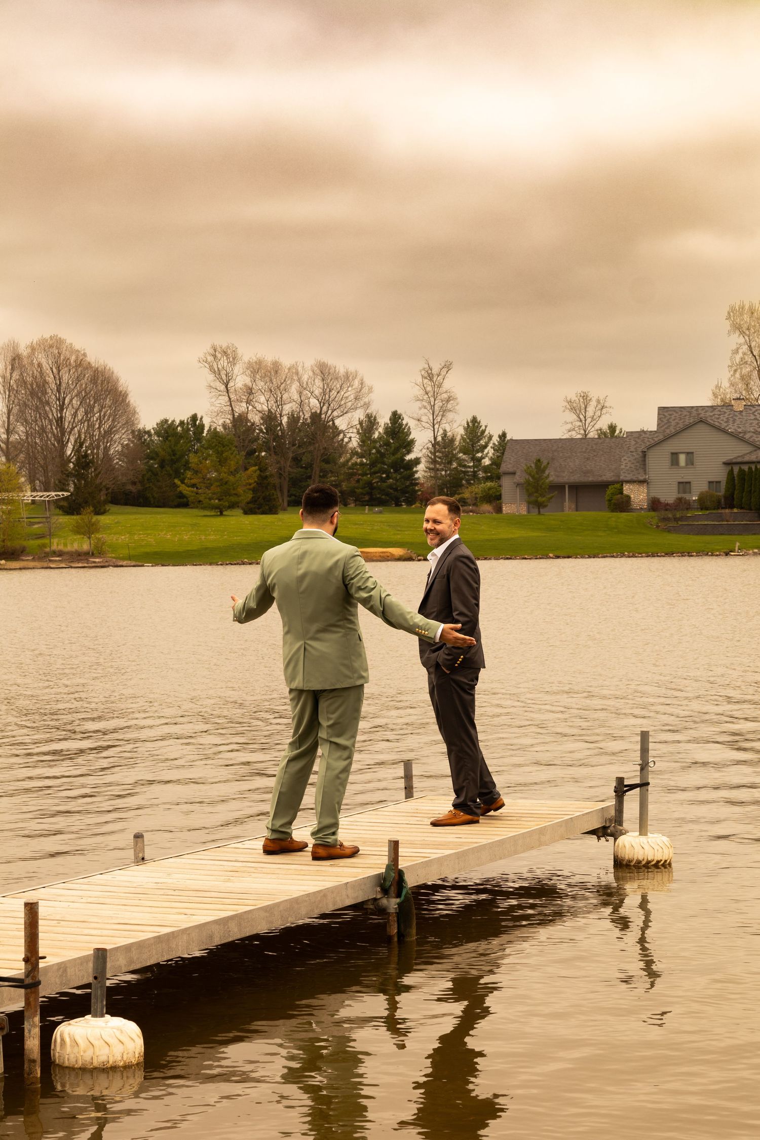 Two grooms in suits stand on a lakeside dock sharing a moment on their wedding day under a dramatic cloudy sky.