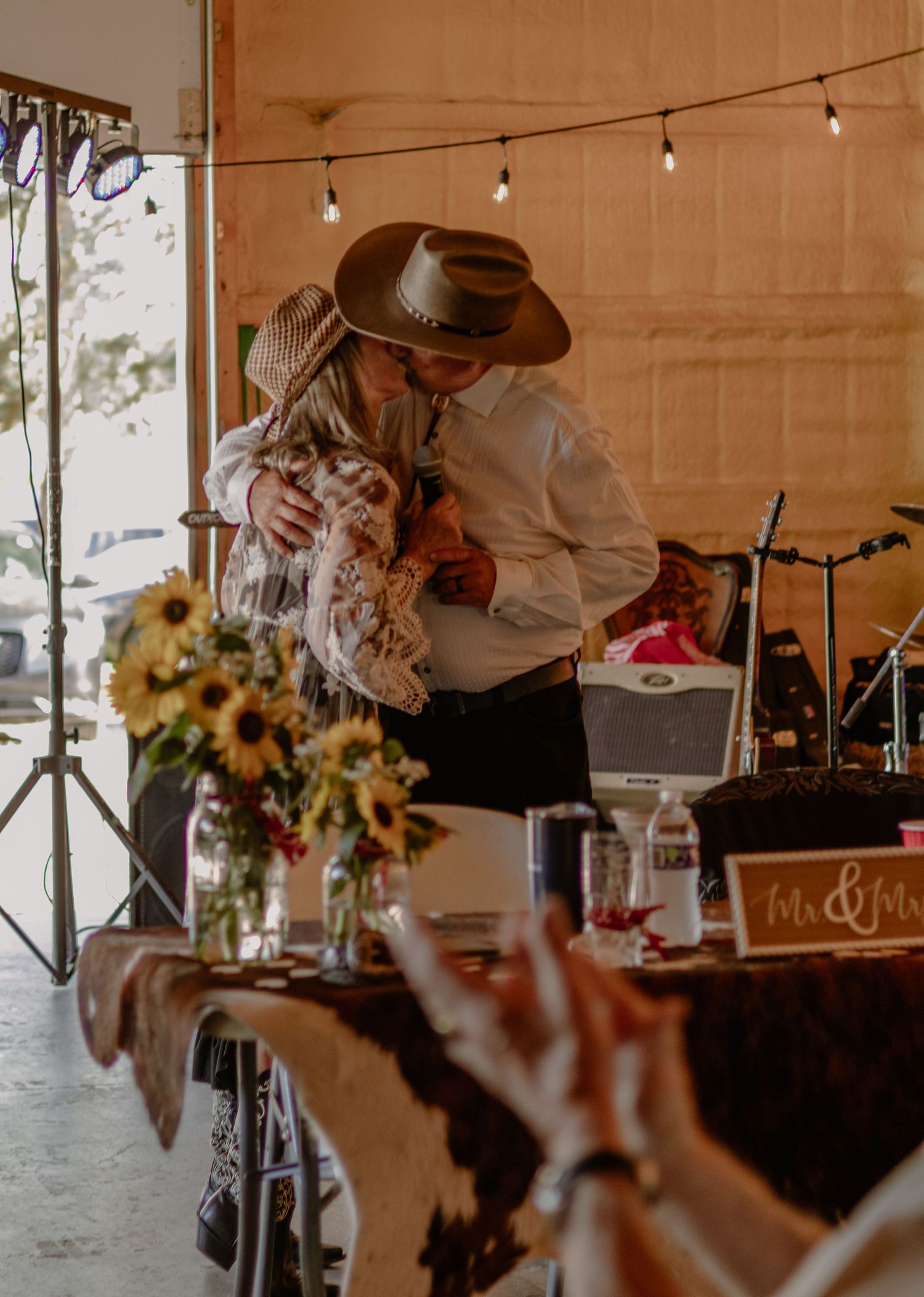 Couple sharing a romantic embrace at a rustic wedding reception with sunflowers and string lights.