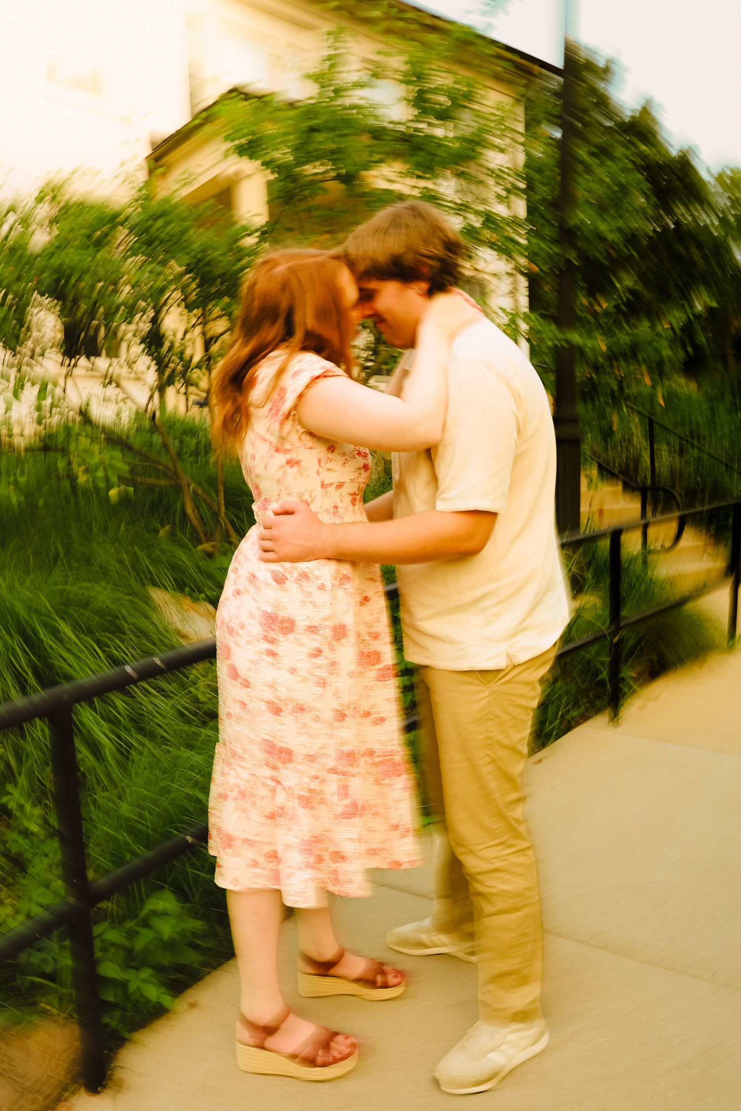 A couple embraces and shares a kiss outdoors on a sidewalk, surrounded by lush greenery, in a dreamy blurred photo.