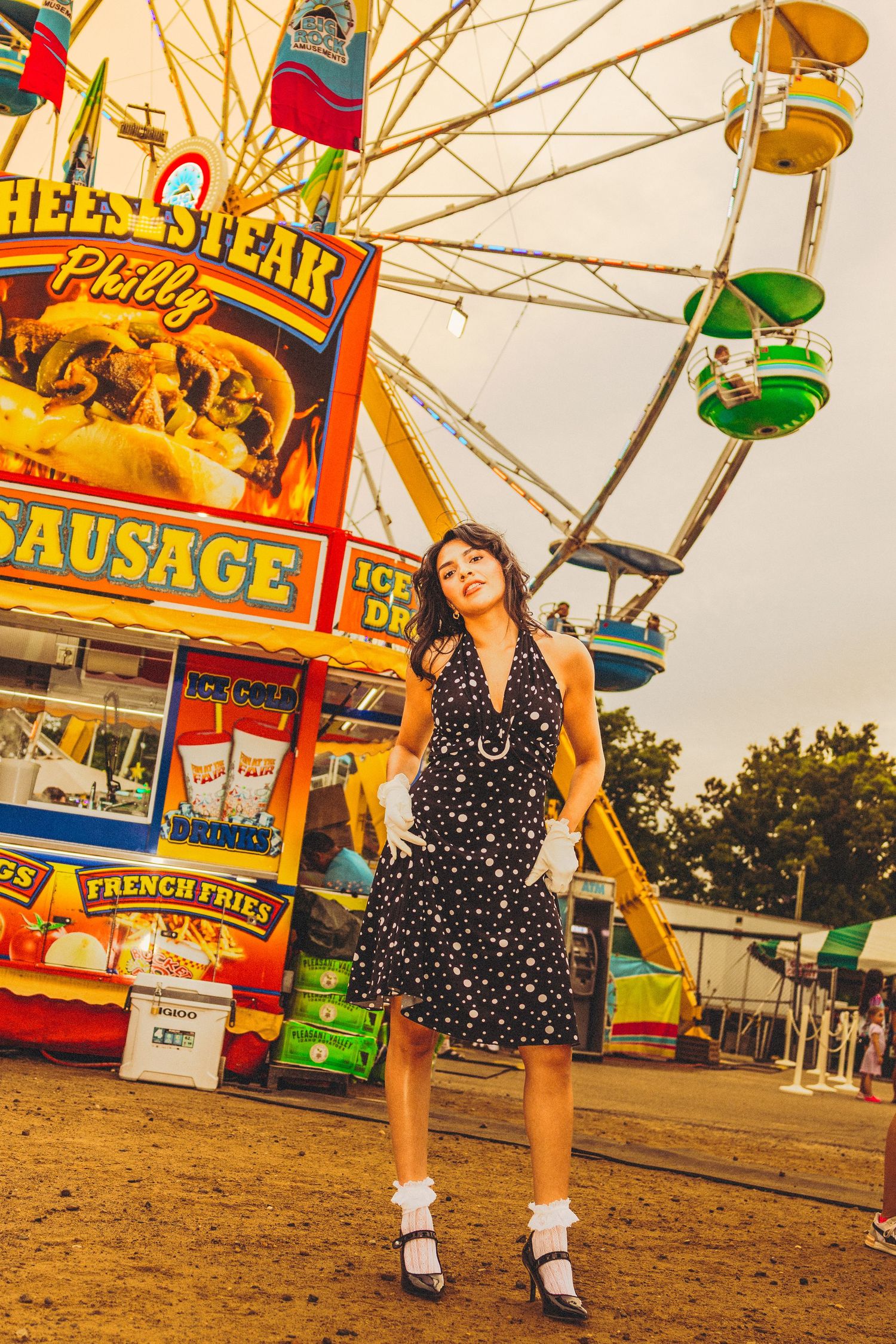 Young woman in polka dot dress posing at a carnival with a Ferris wheel and food stand in the background.