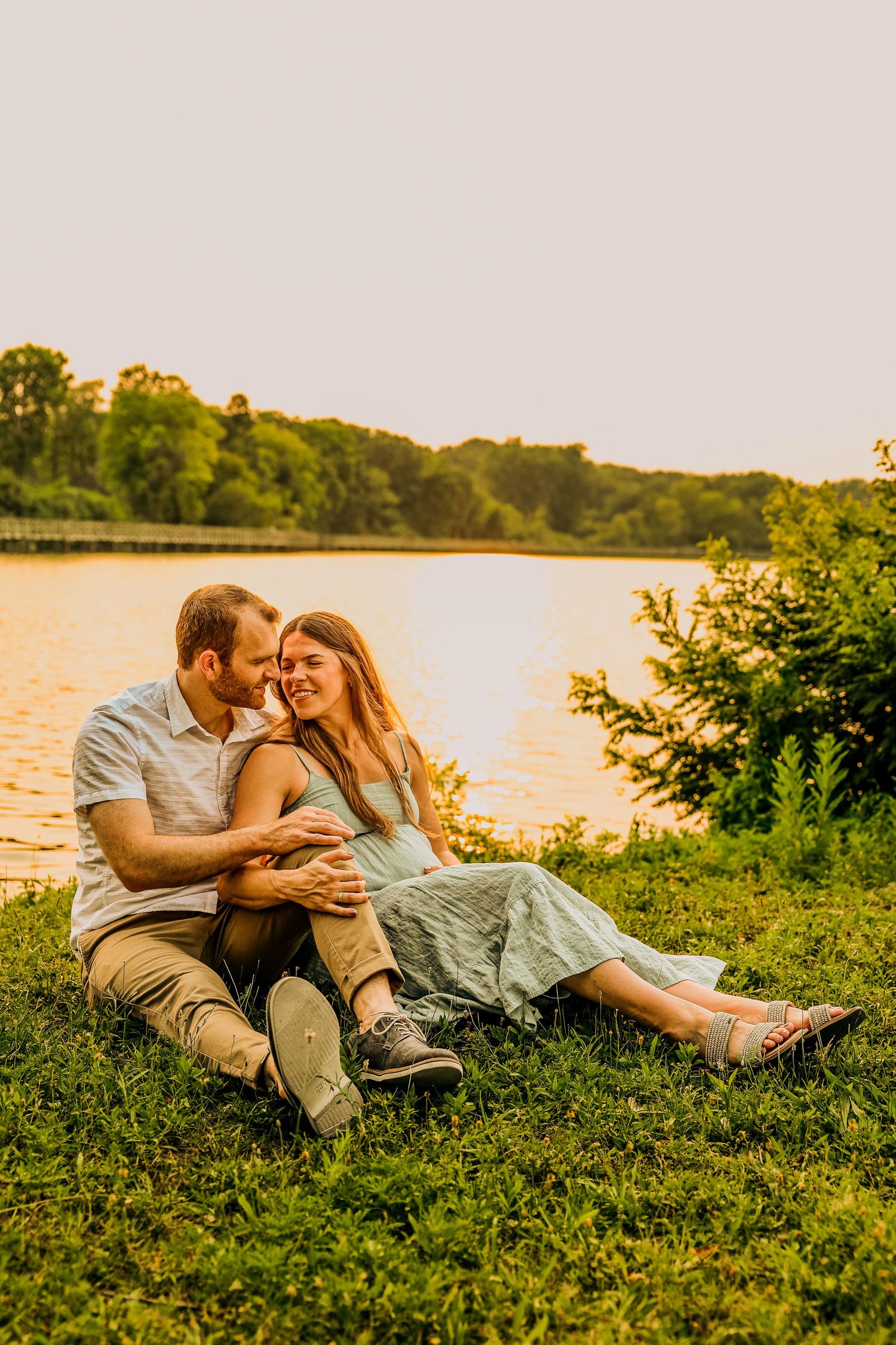 Couple sitting on grass by a golden sunset lake, man embracing woman in mint dress, romantic outdoor engagement photo.
