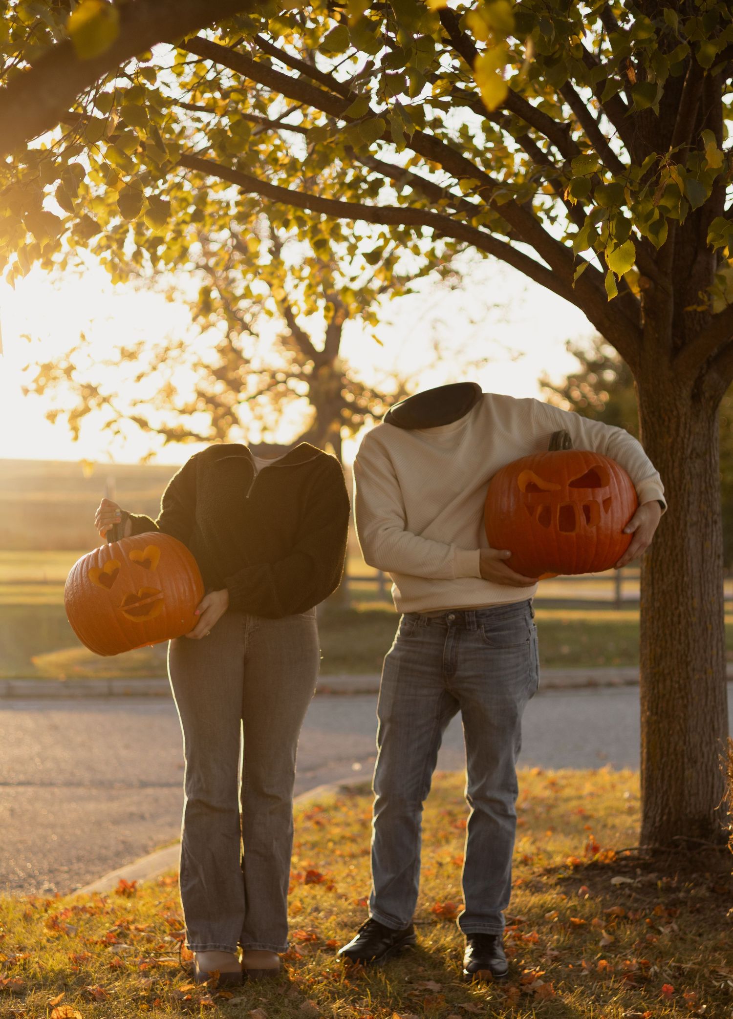 Two people holding carved jack-o-lanterns over their faces under a tree during golden hour in autumn.