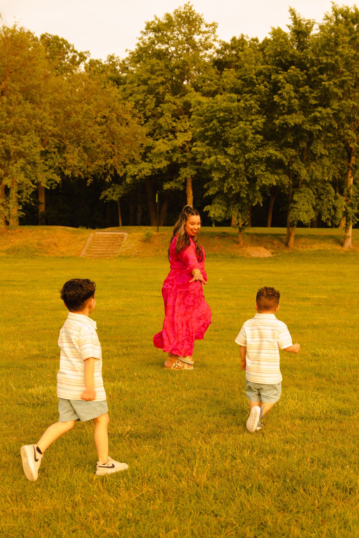 Two young boys run toward their mother in a red dress on a sunny green lawn surrounded by trees.