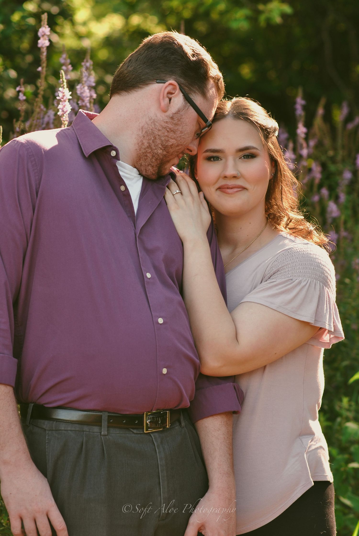 Romantic couple embraces in a garden setting with purple flowering trees in the background.