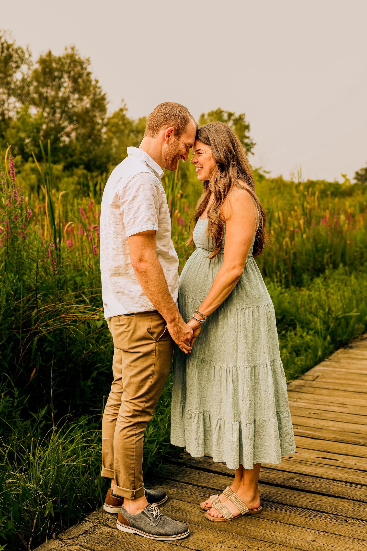 Couple holding hands on a wooden boardwalk surrounded by lush greenery, woman in green dress showing baby bump.