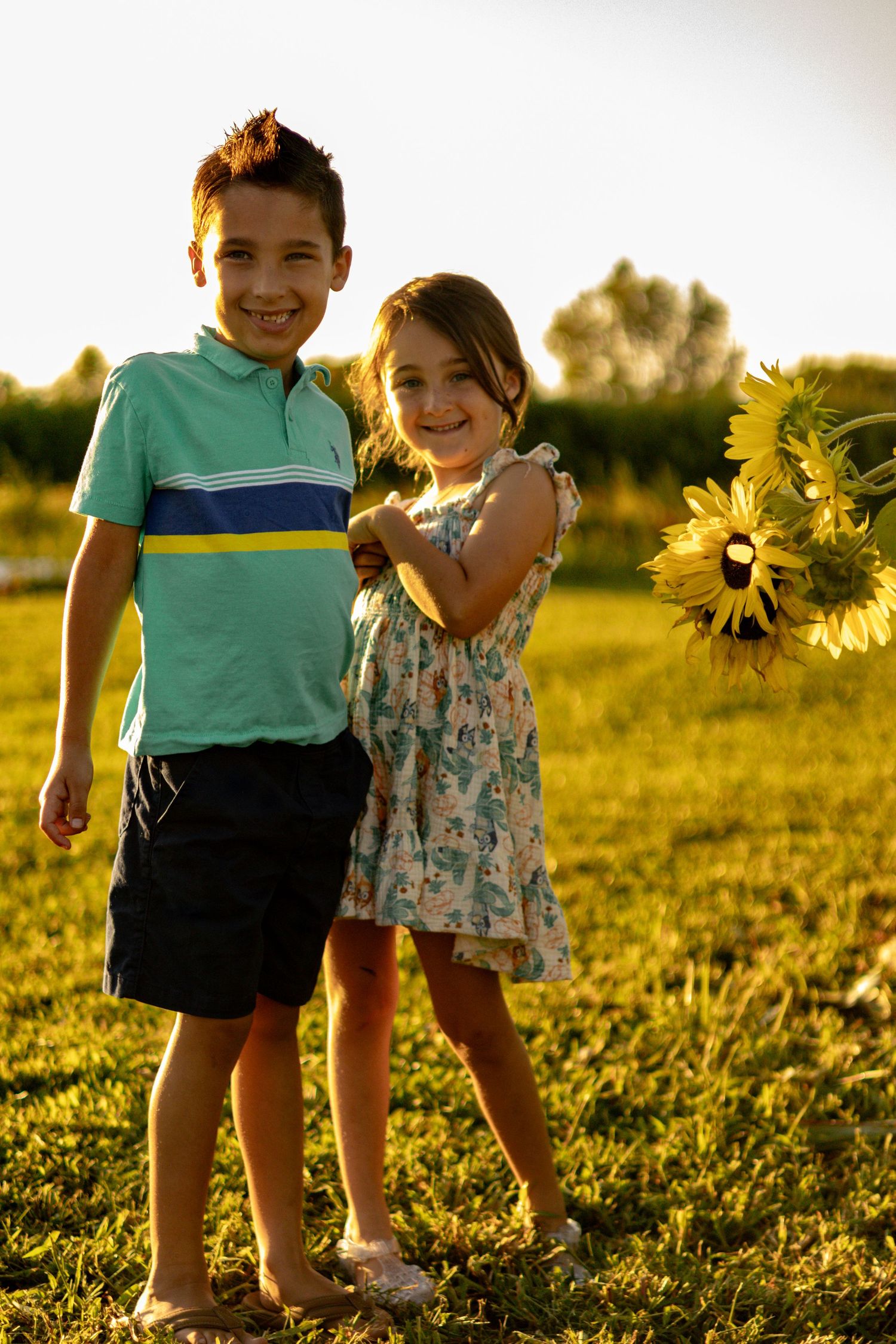 Two young children stand together in a sunlit field of sunflowers during golden hour.