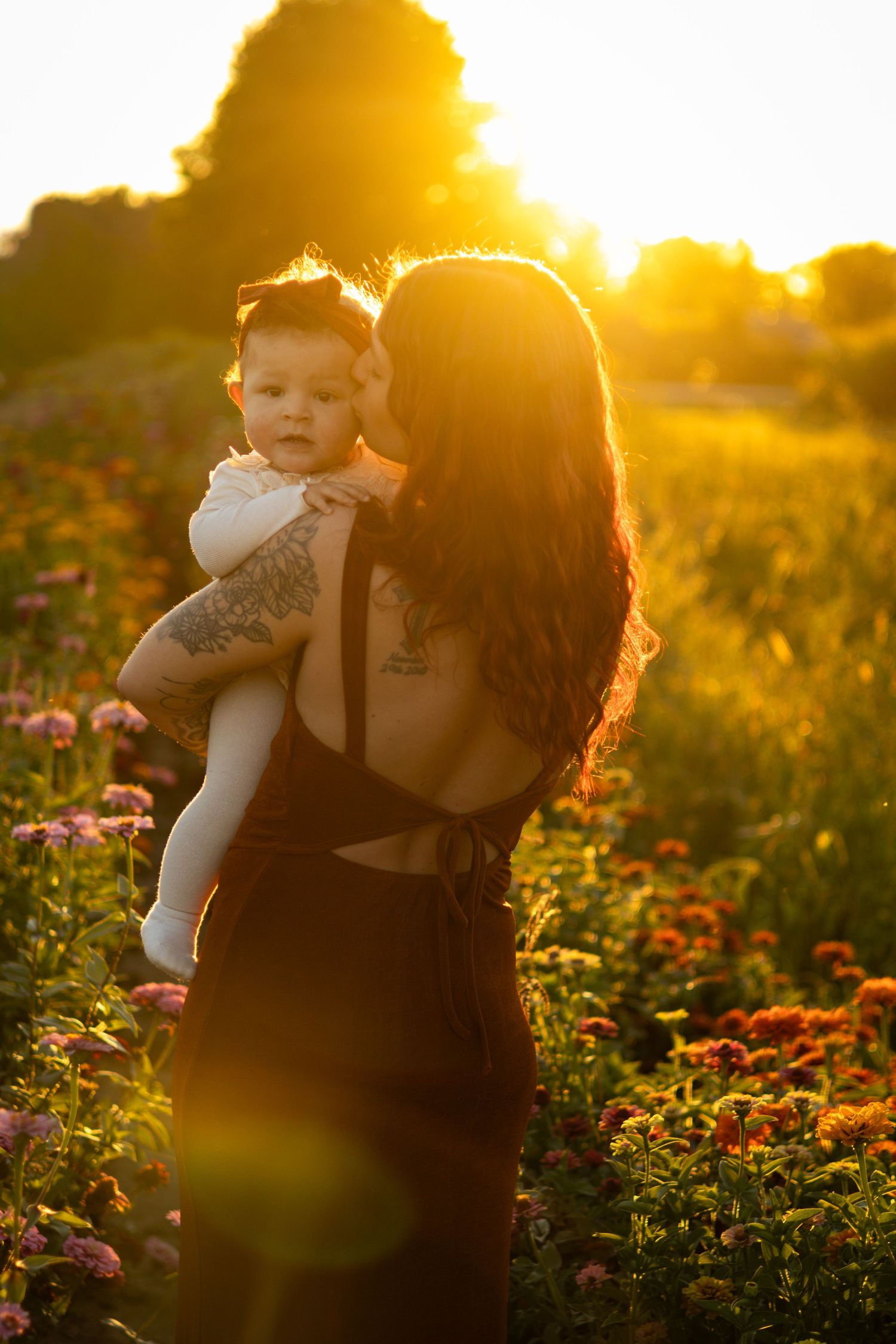 A tattooed mother holds her baby in a sunlit flower field at golden hour, surrounded by colorful blooms.