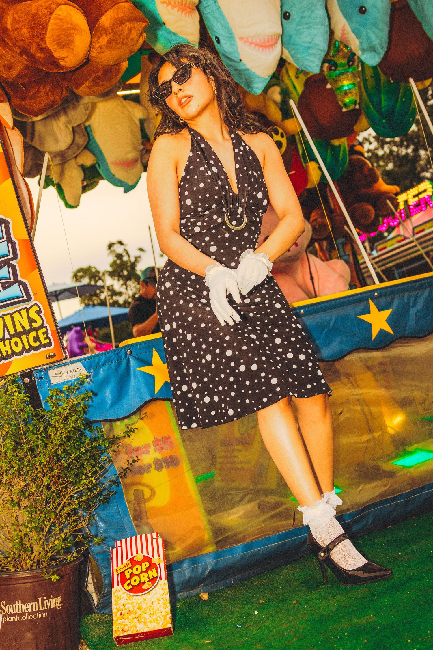 Woman in polka dot halter dress and white gloves posing at a colorful carnival game booth with stuffed animal prizes.