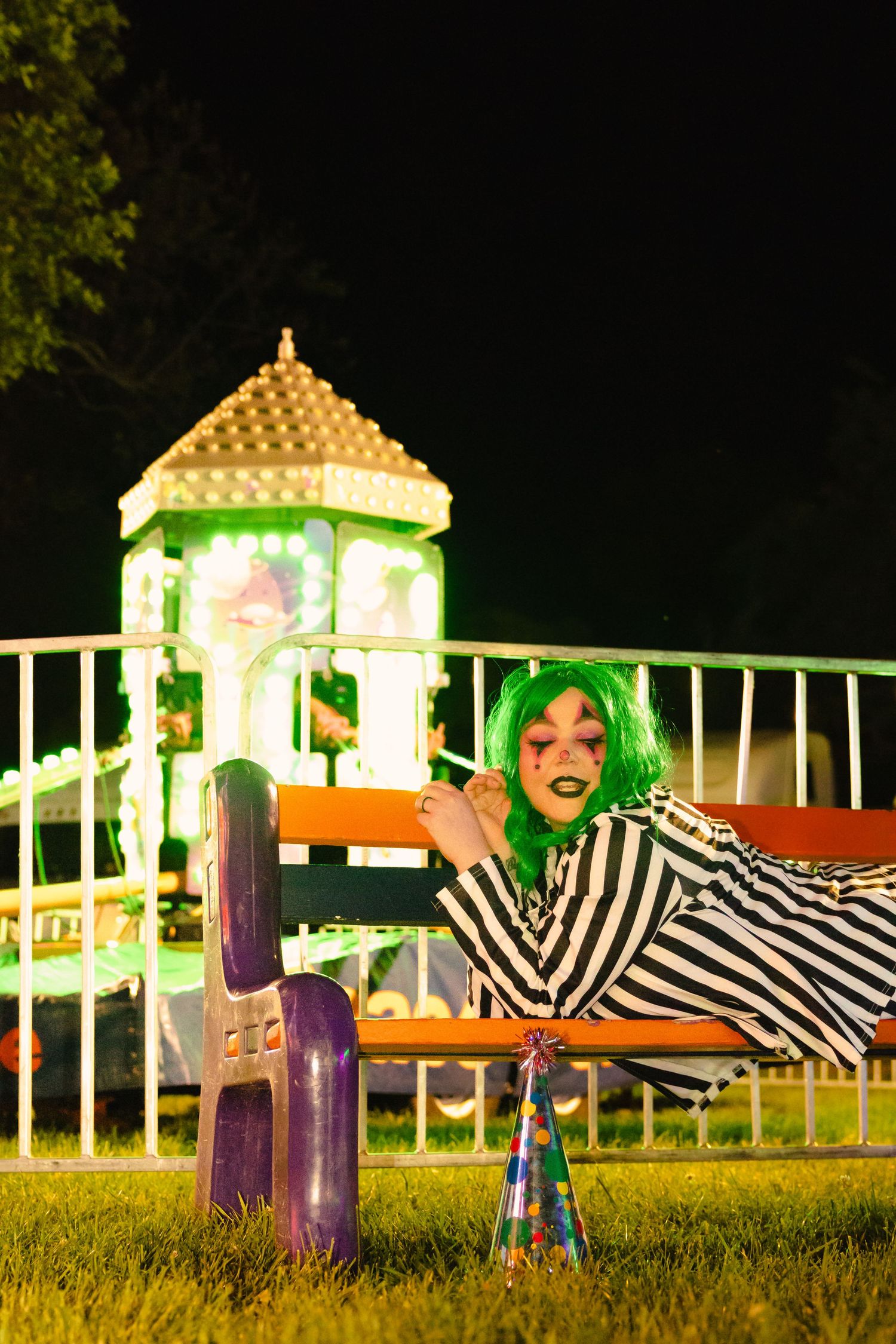 Person in clown costume relaxing on bench at illuminated fairground attraction at night.