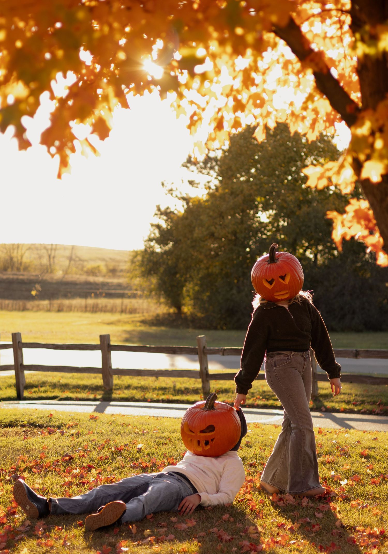 Two children with jack-o-lantern heads pose in a fall yard with autumn leaves and a wooden fence backdrop.