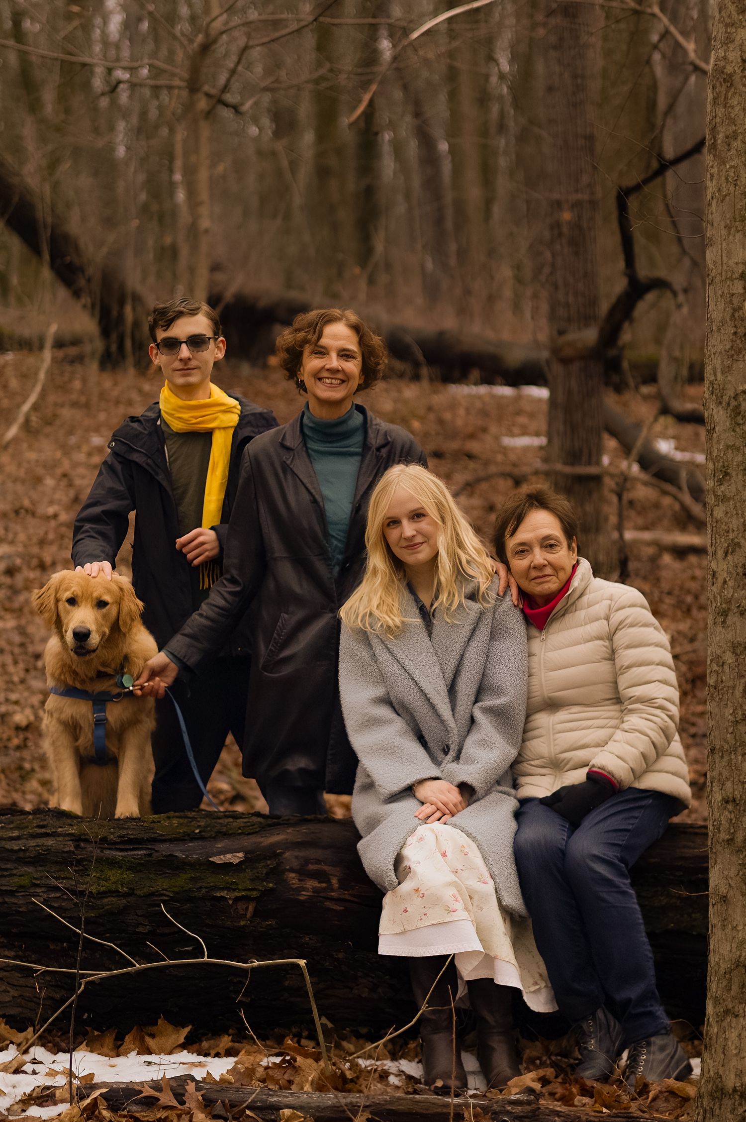Family of four poses with golden retriever on fallen log in autumn forest, wearing cozy coats and scarves.